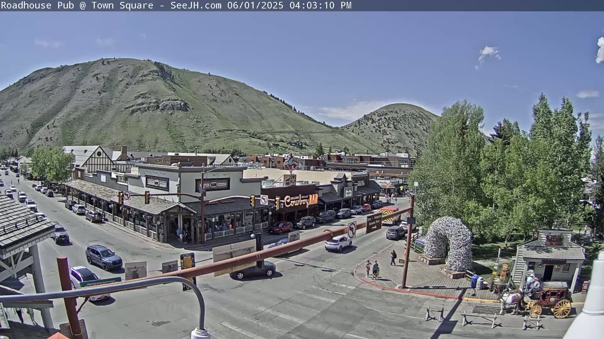 A sunny daytime view of a town square nestled between mountains, with cars parked along the streets and a horse-drawn carriage near a small building.