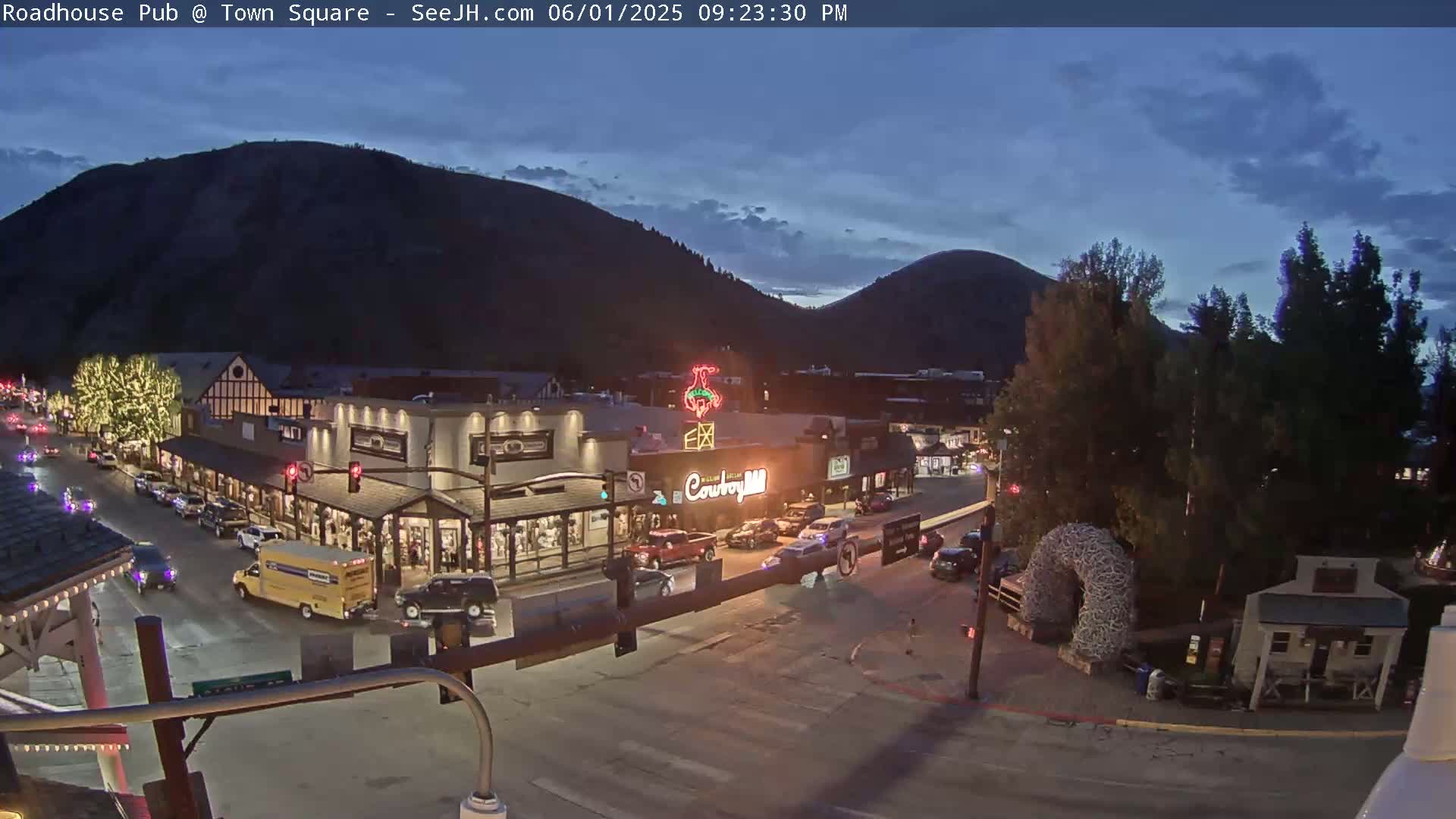 A nighttime view of a town square with illuminated buildings, cars on the street, and mountains in the background under a twilight sky.