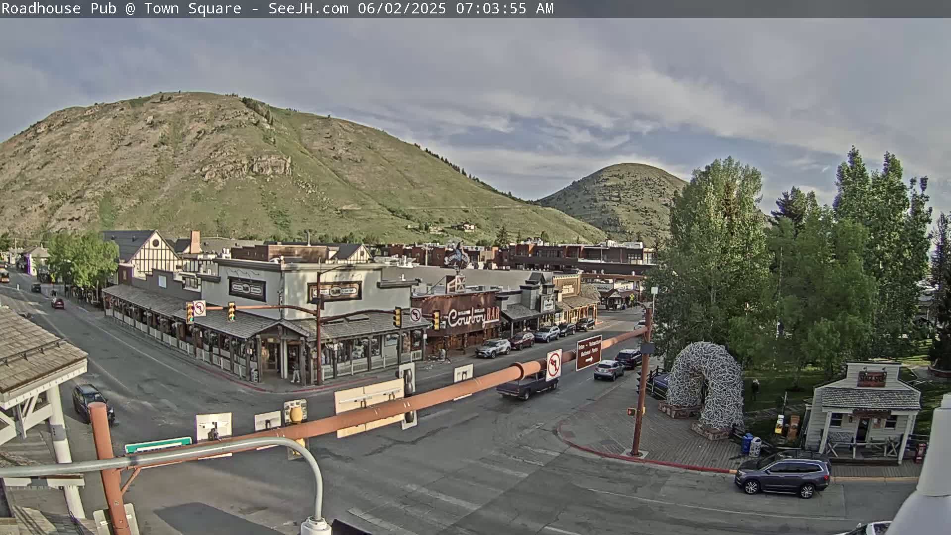 A partly cloudy morning reveals a mountain town square with several shops and cars parked along the streets, nestled between two hills.