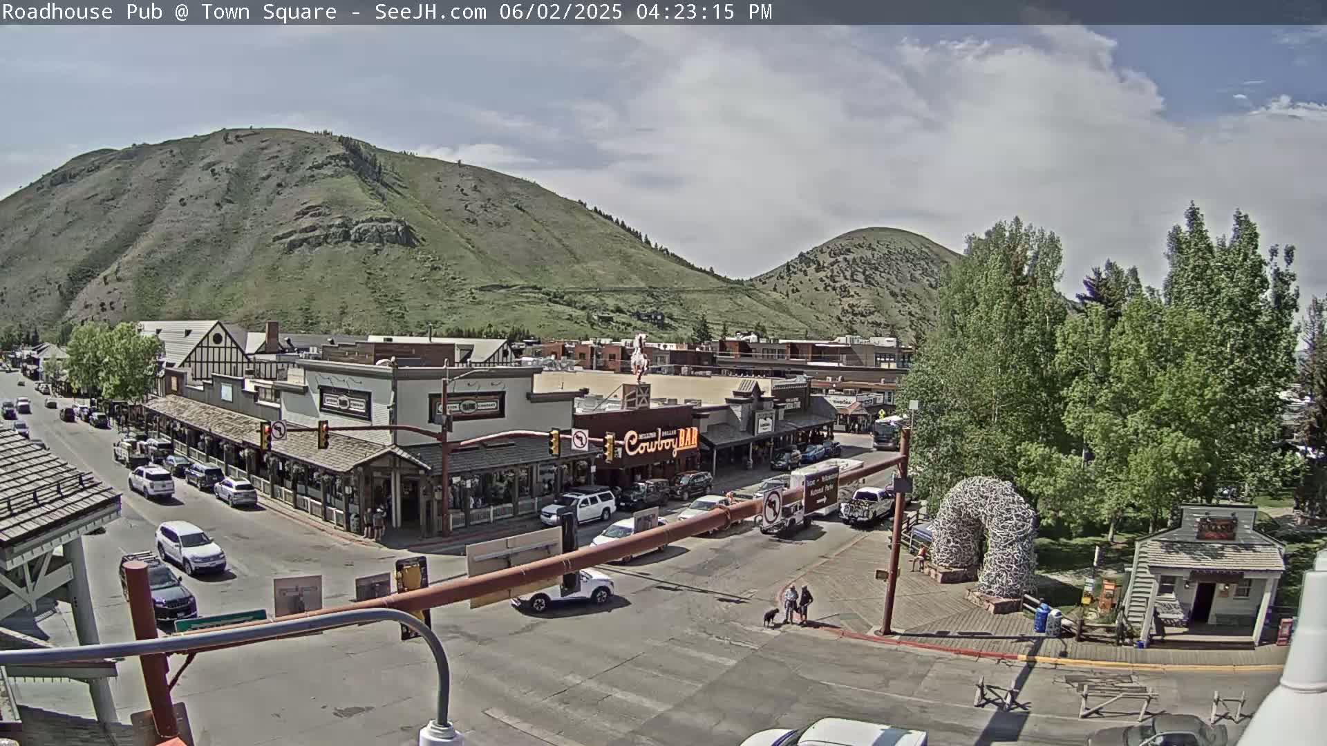 A partly cloudy day reveals a town square nestled between two hills, with cars parked along the streets and buildings lining the square.