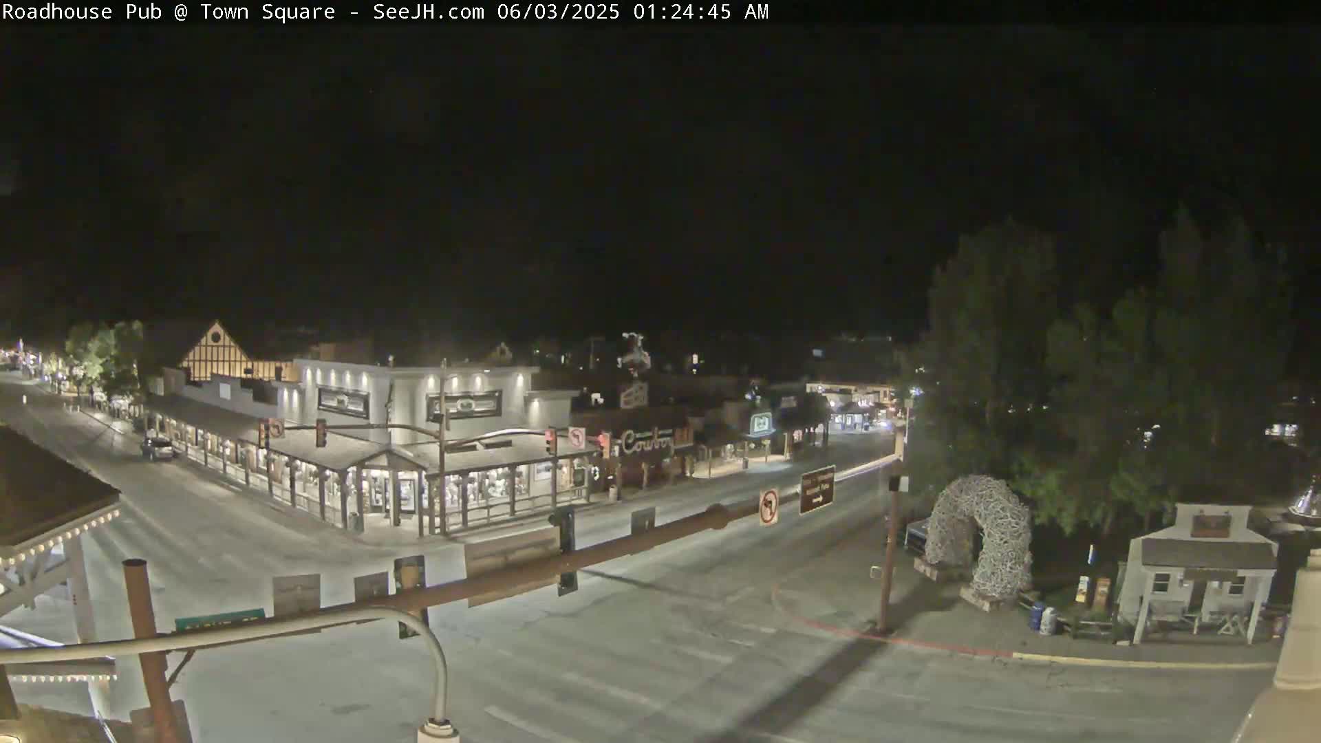 A nighttime, snow-covered town square with several buildings and a large, arched sculpture.