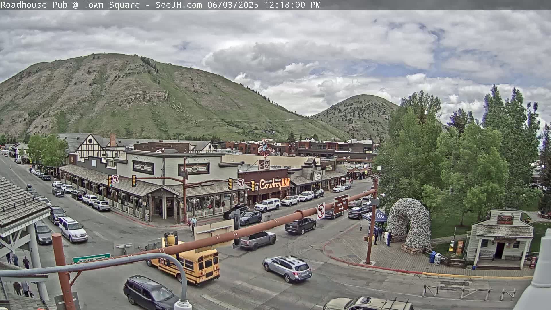 A partly cloudy day reveals a mountain town square with buildings, cars, and a school bus, all surrounded by green hills.