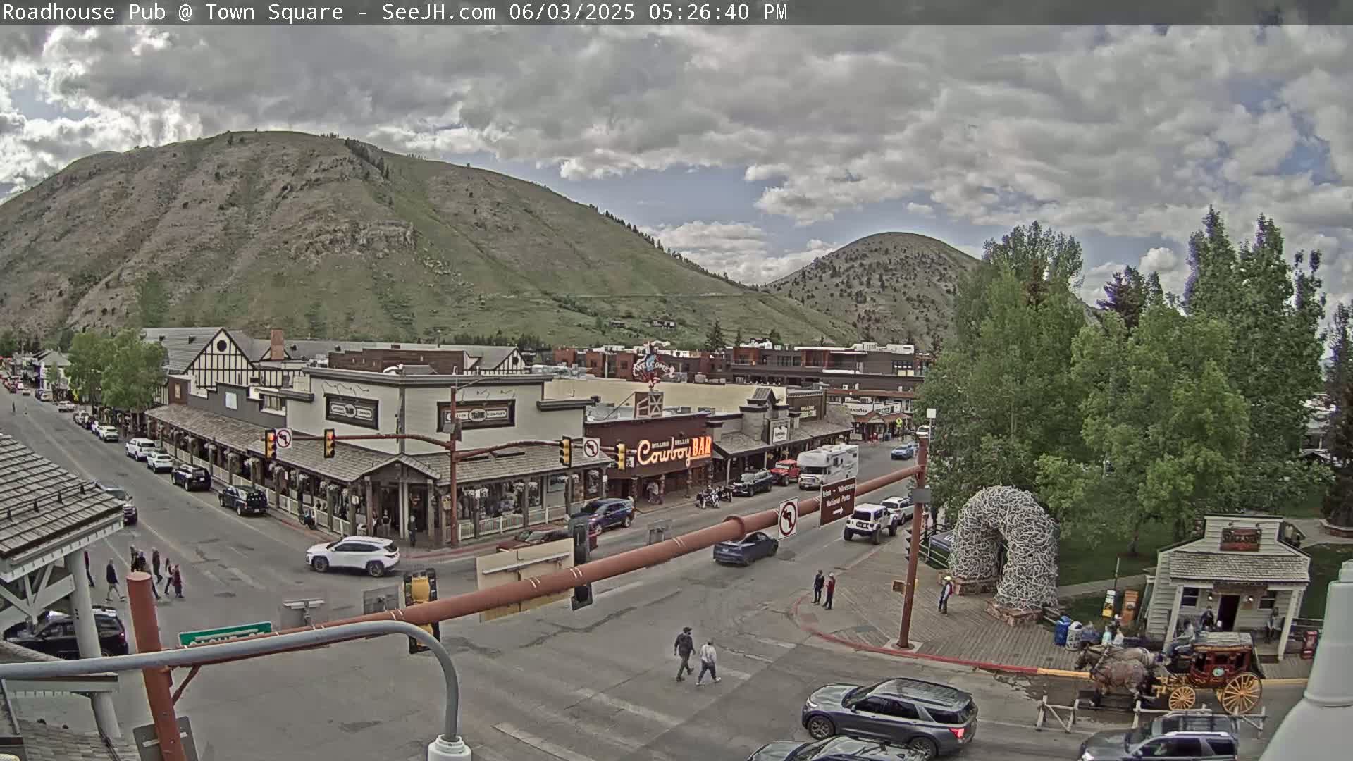 A partly cloudy day reveals a mountain town square with buildings, vehicles, and pedestrians, under a mostly cloudy sky.