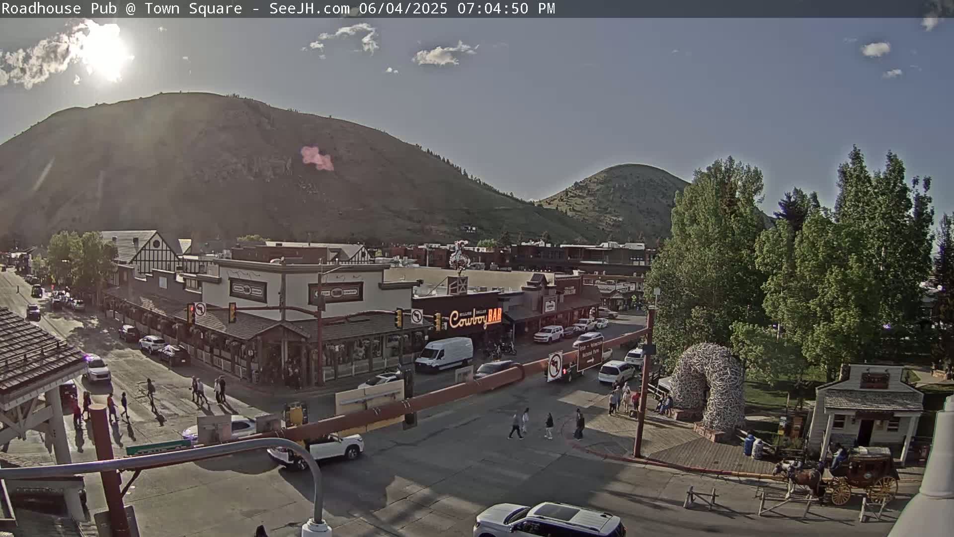 A high-angle, daytime view of a town square nestled between hills under mostly sunny skies, showing pedestrians, vehicles, and buildings, including a saloon, with a horse-drawn carriage present near a large antler arch.