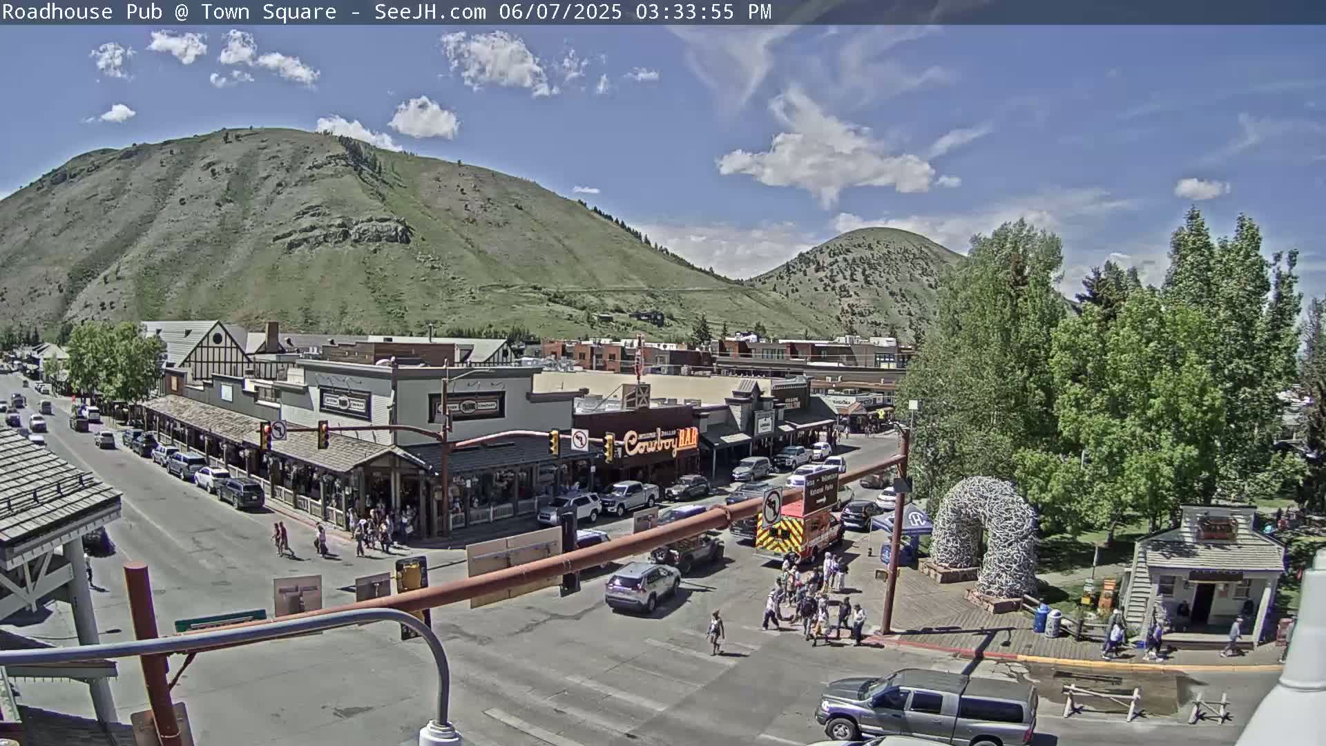 A partly sunny day reveals a town square with shops and pedestrians, nestled at the base of a large, green mountain.