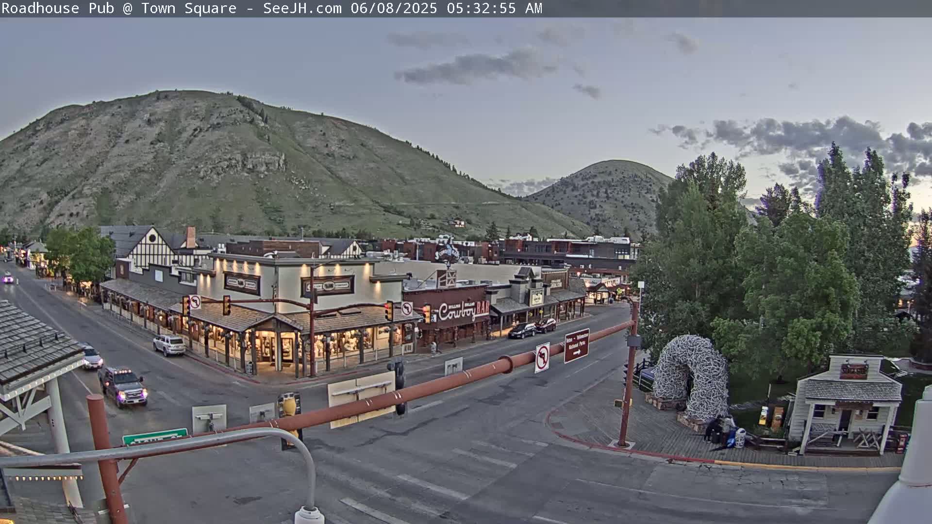 A town square nestled between hills under a partly cloudy twilight sky, showing several buildings, vehicles, and a large antler arch.