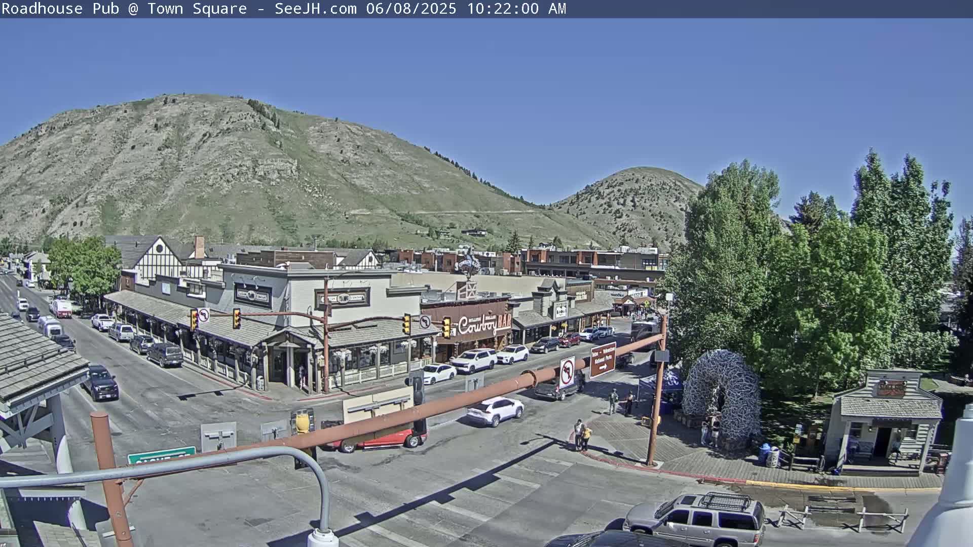 A sunny day reveals a town square nestled between hills, featuring several buildings, parked cars, and pedestrians near a crossroads.
