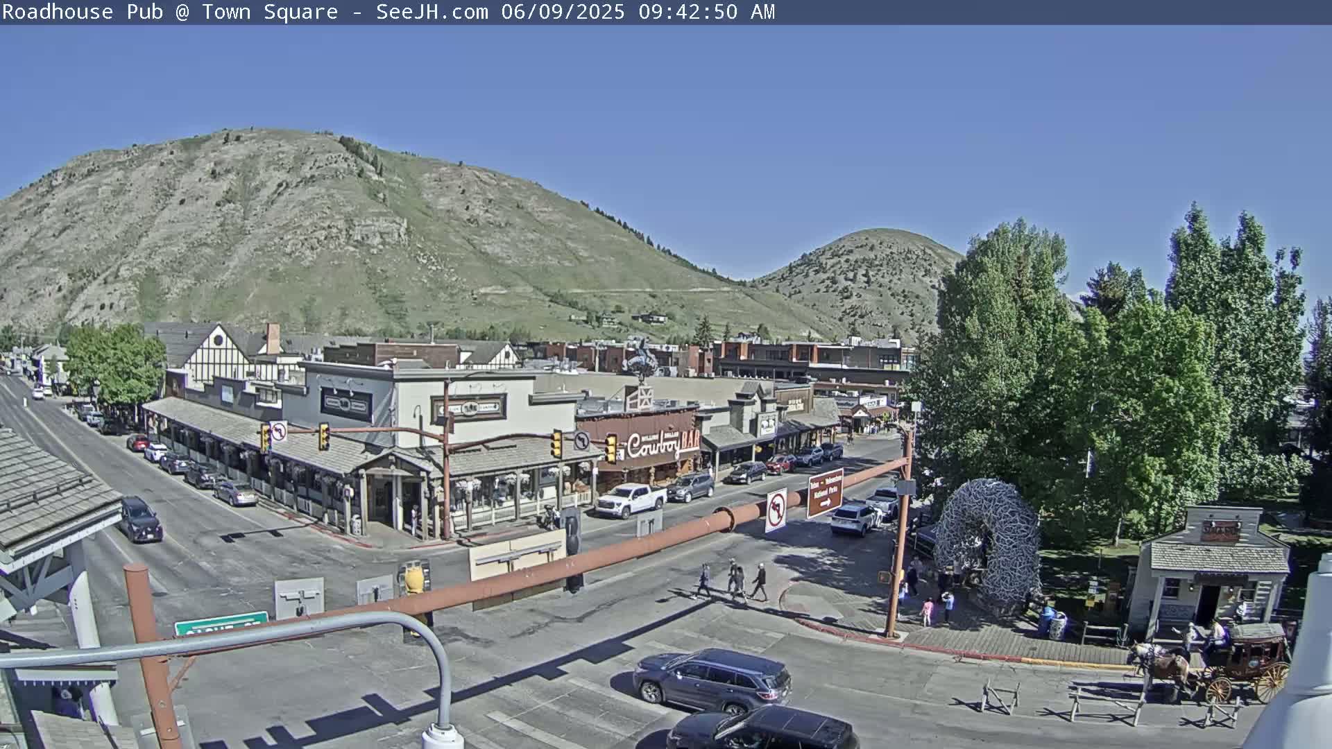 A sunny day reveals a town square with buildings, vehicles, and pedestrians, nestled between two hills.