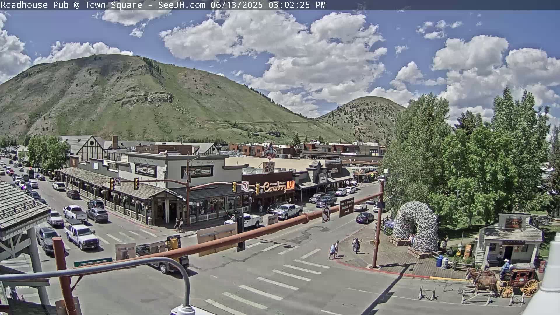 A partly cloudy day reveals a mountainside town square with vehicles on the streets, several buildings, and a horse-drawn carriage.
