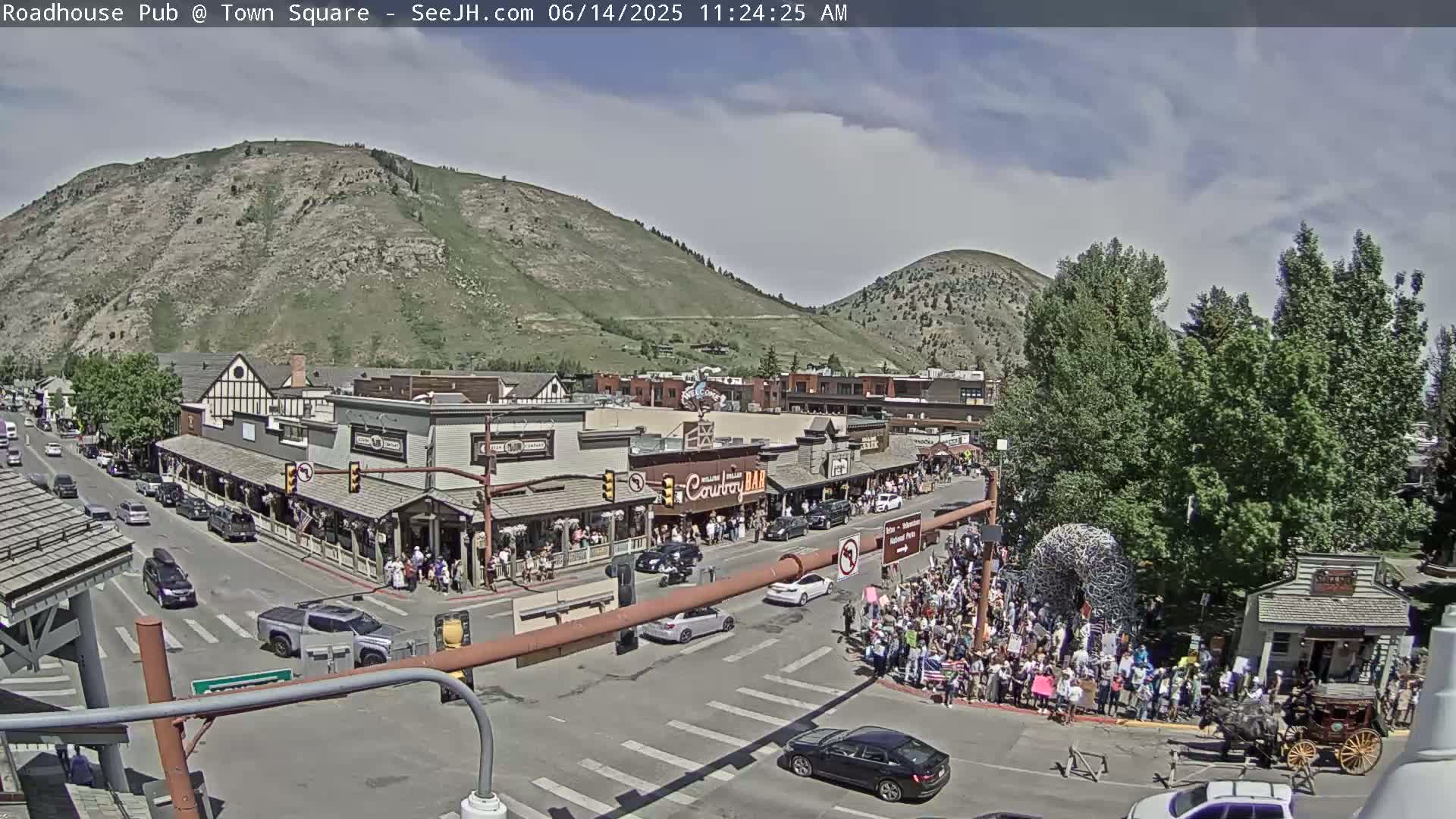 A partly sunny day reveals a town square with many people gathered, cars driving on the streets, and buildings lining the sides, all nestled at the base of a large, green mountain.