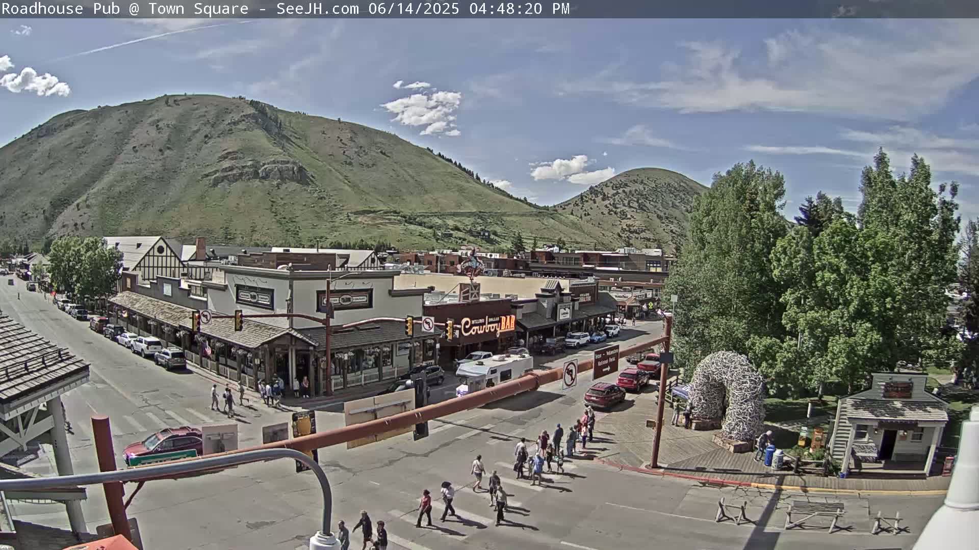 A partly sunny day reveals a town square with many people gathered, cars driving on the streets, and buildings lining the sides, all nestled at the base of a large, green mountain.