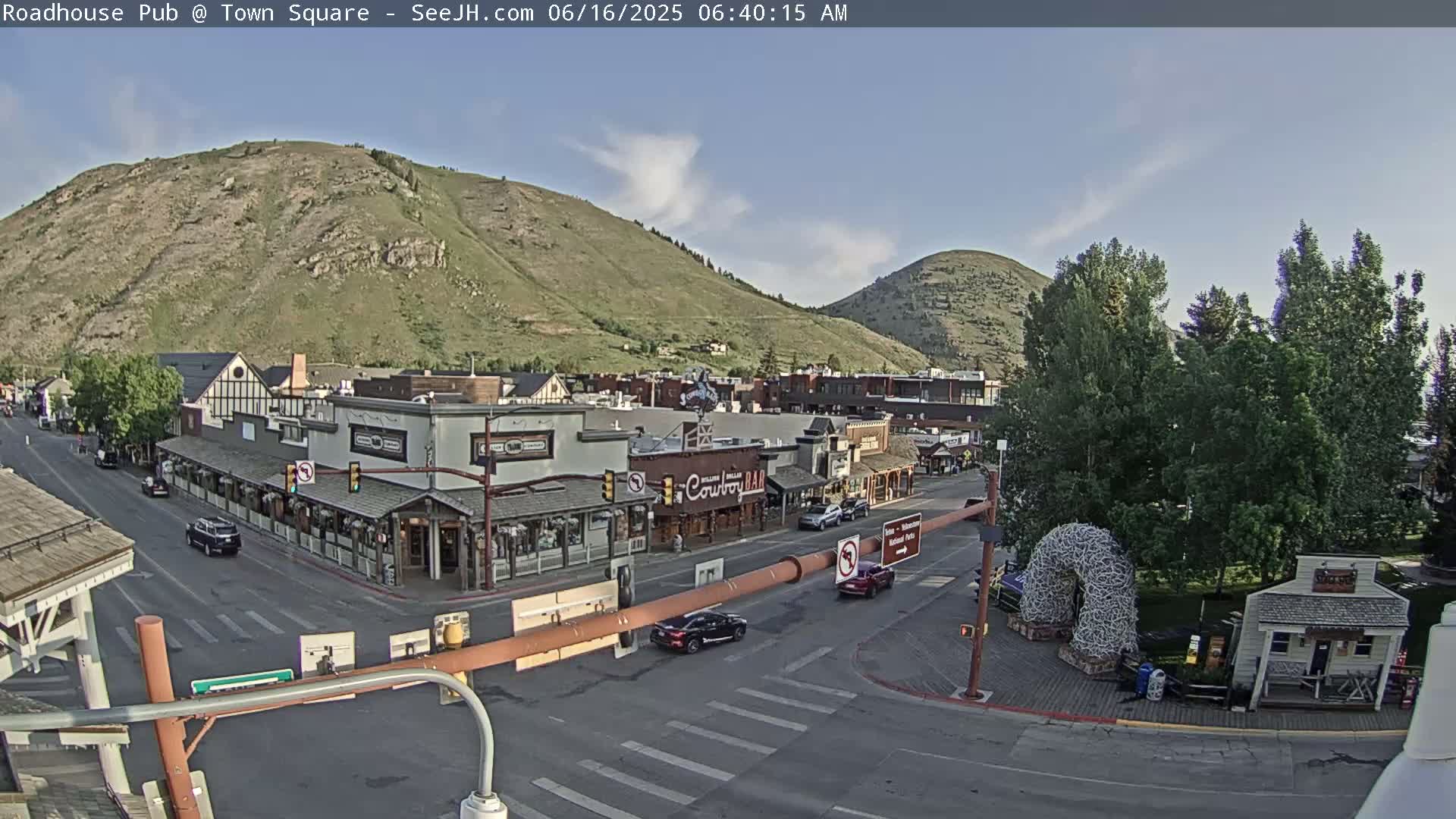 A partly sunny view of a town square nestled between mountains, with cars parked along the street and several buildings lining the square.