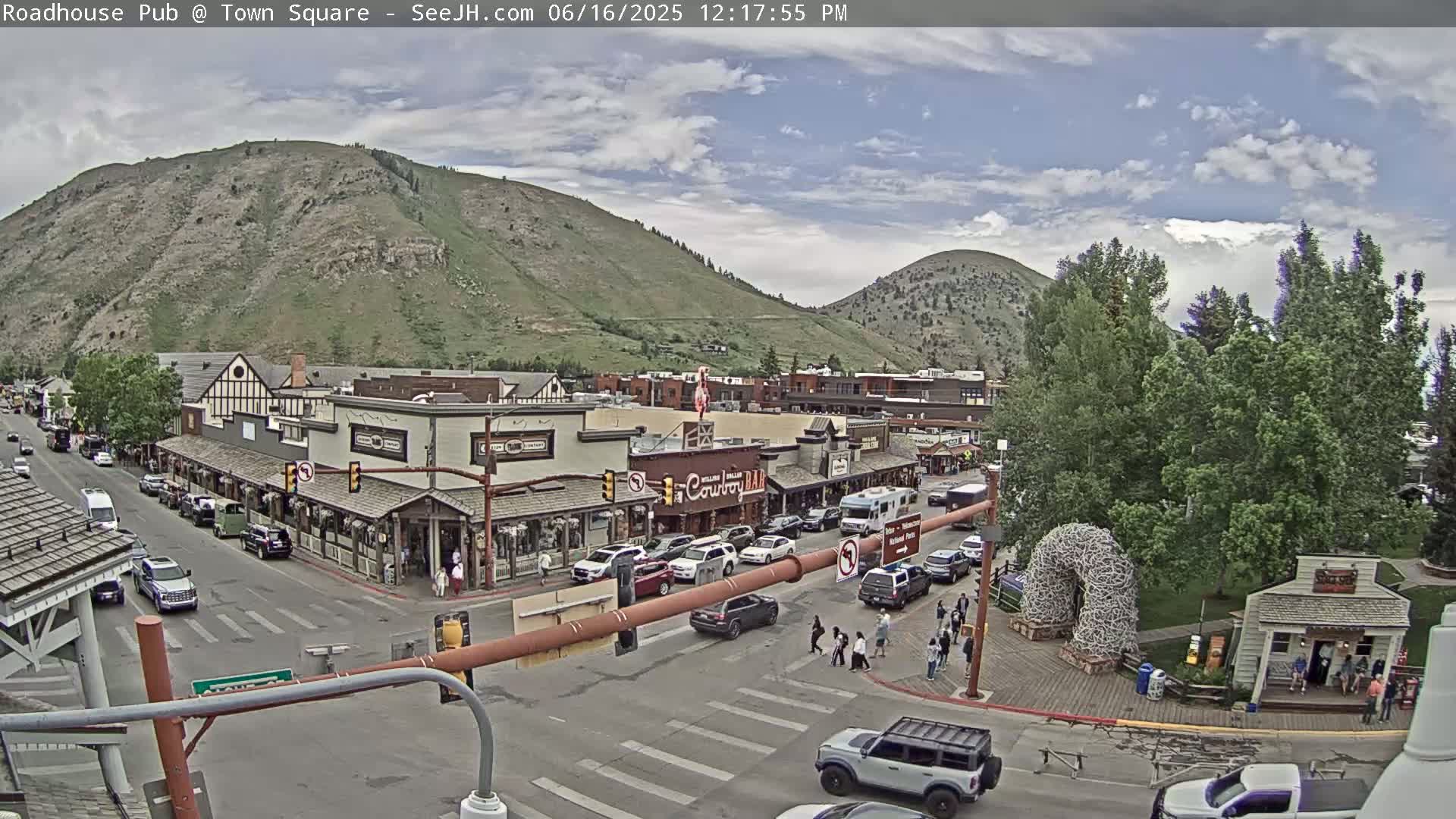 A partly cloudy day shows a town square with several buildings, cars, and pedestrians, nestled at the base of a mountain range.
