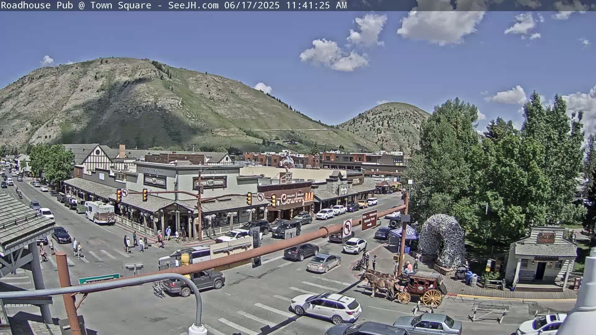 A sunny day in a town situated in a valley between mountains shows a street scene with cars, pedestrians, a horse-drawn carriage, and numerous shops and buildings.