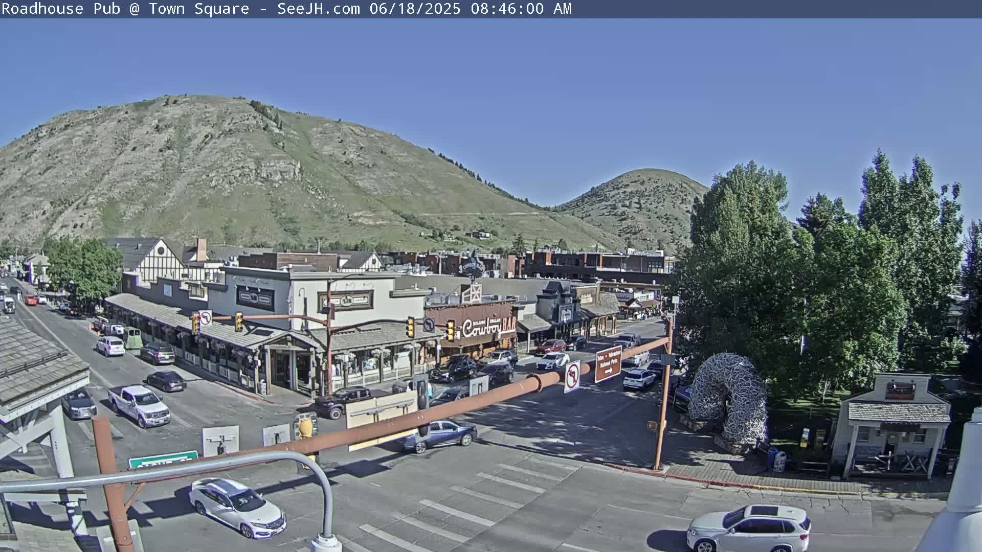 A sunny day reveals a town square with cars driving on the streets, buildings lining the streets, and mountains in the background.