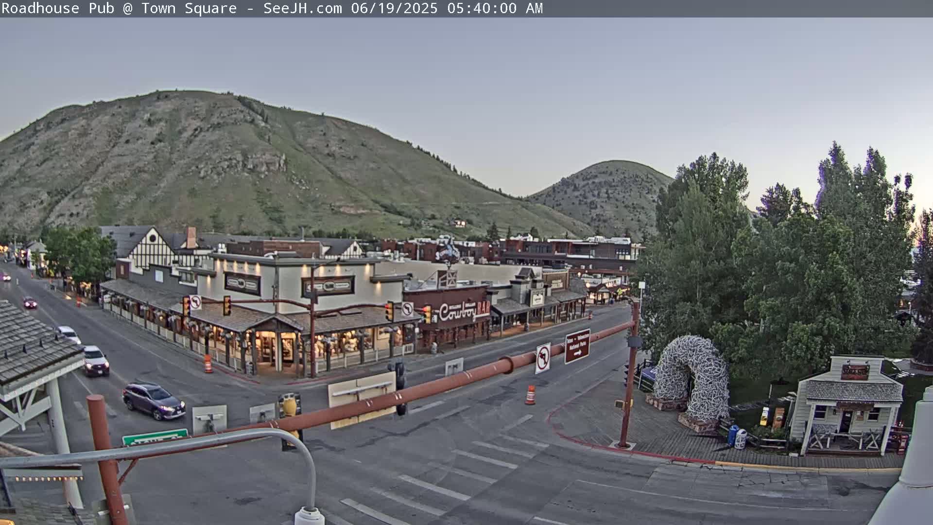 A mostly clear dawn illuminates a town square nestled between hills, with several cars on the street and some pedestrians visible near shops and restaurants.