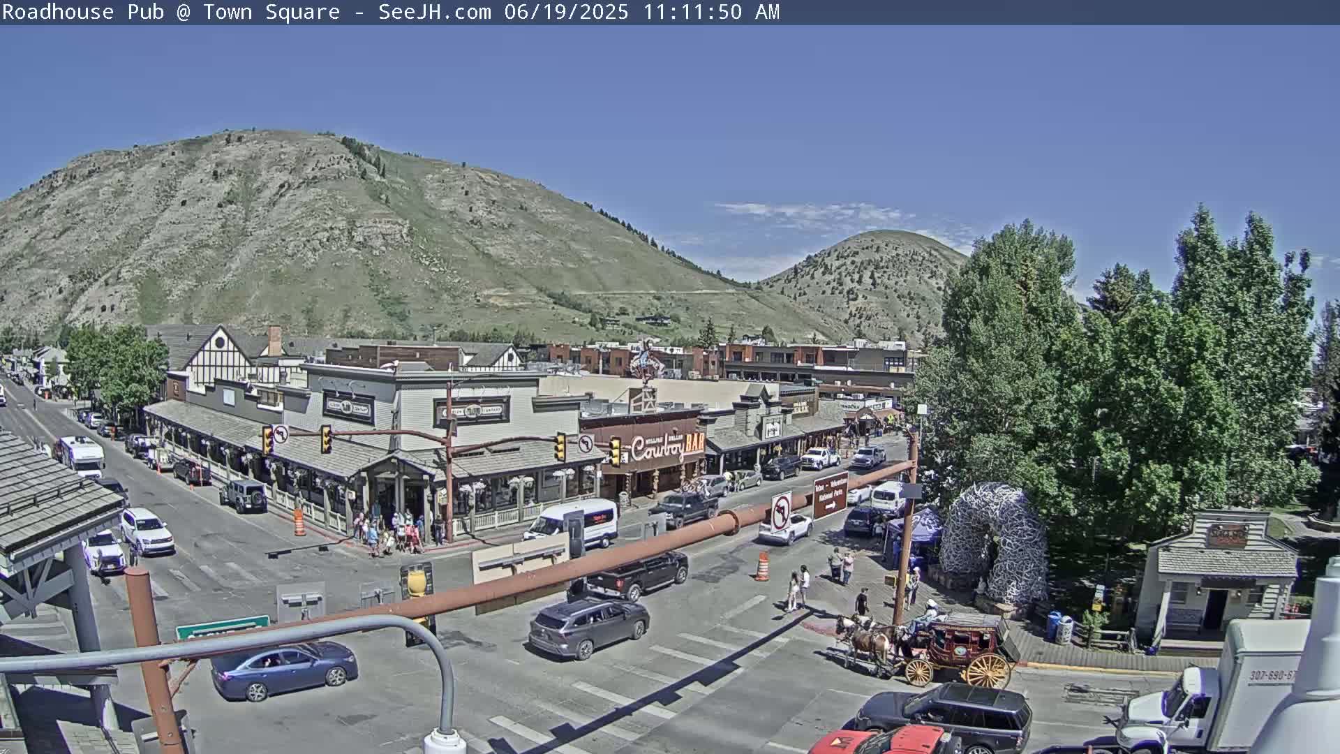 A sunny day reveals a town square with buildings, vehicles, and pedestrians, situated at the base of a mountain.