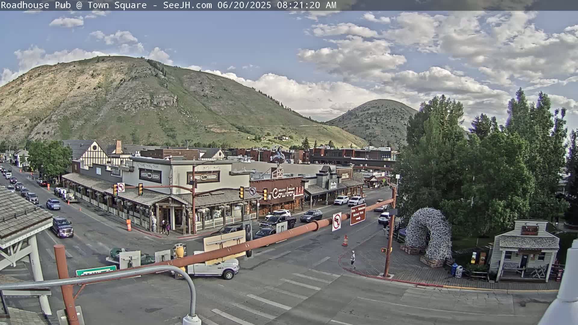 A partly cloudy day reveals a town square nestled at the base of a mountain, with cars parked along the streets and buildings lining the square.