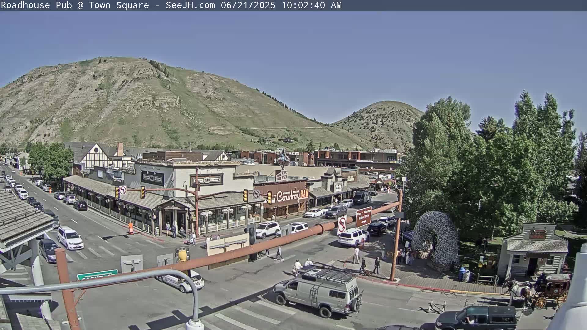 A sunny day reveals a town square nestled between mountains, with cars parked along streets and people walking around.