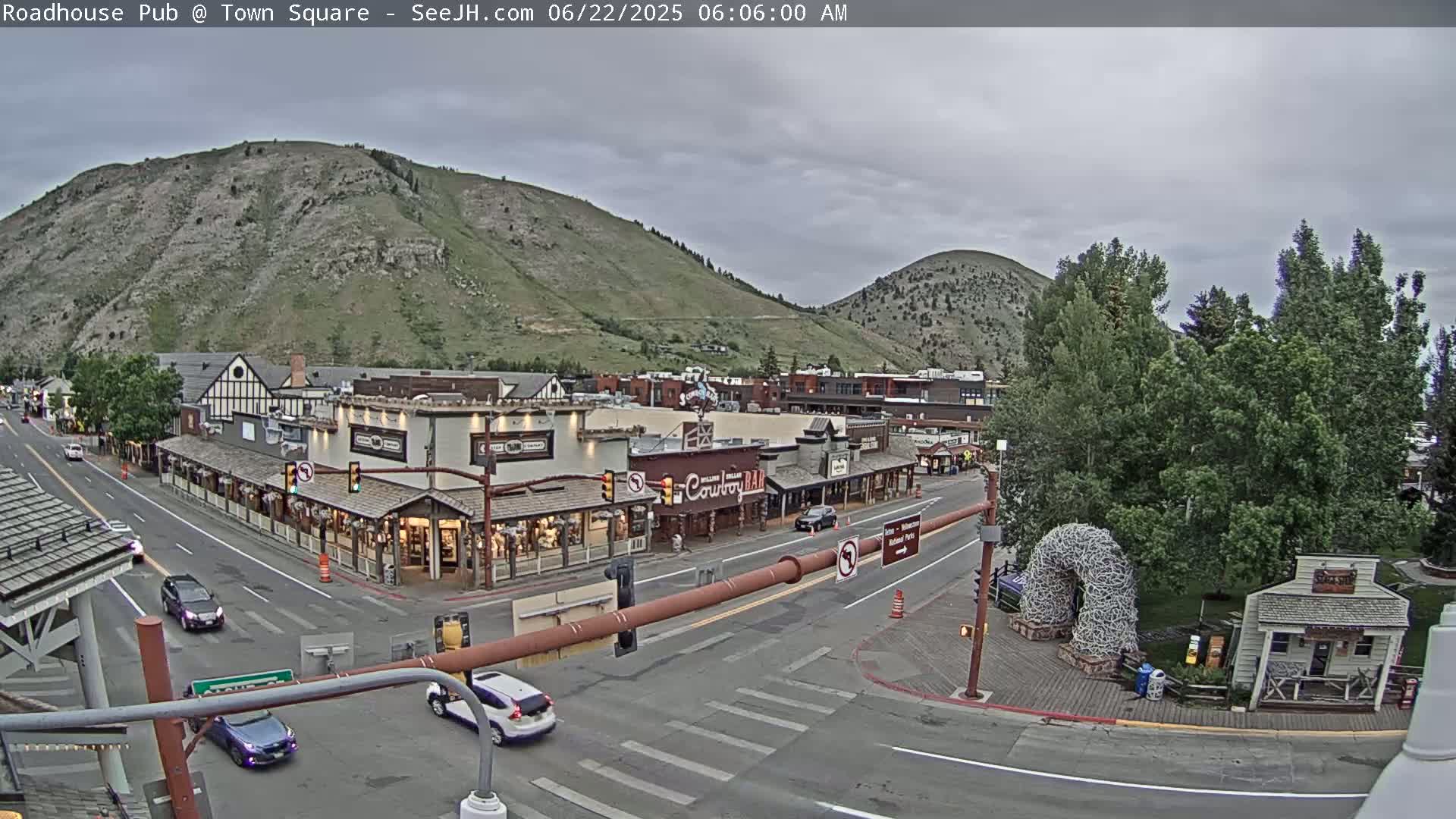 A mountain town square with overcast skies, featuring several buildings, vehicles on the road, and a large antler arch.