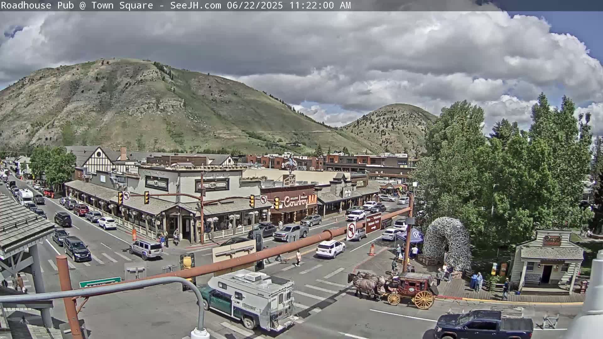A partly cloudy day reveals a town square nestled beneath a mountain, with a horse-drawn carriage and numerous vehicles parked and driving along the streets.