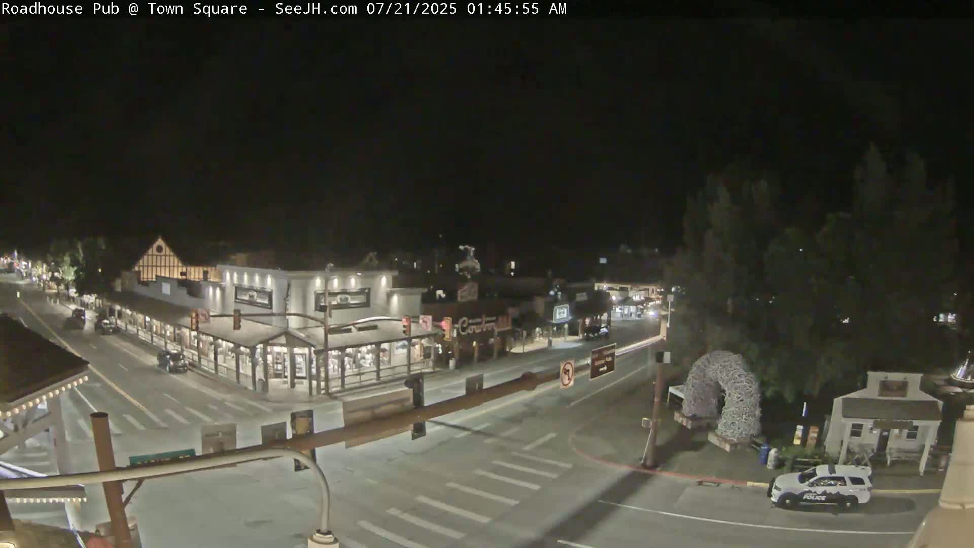 A nighttime overhead view of a town square with a few cars and a police vehicle, under clear skies.