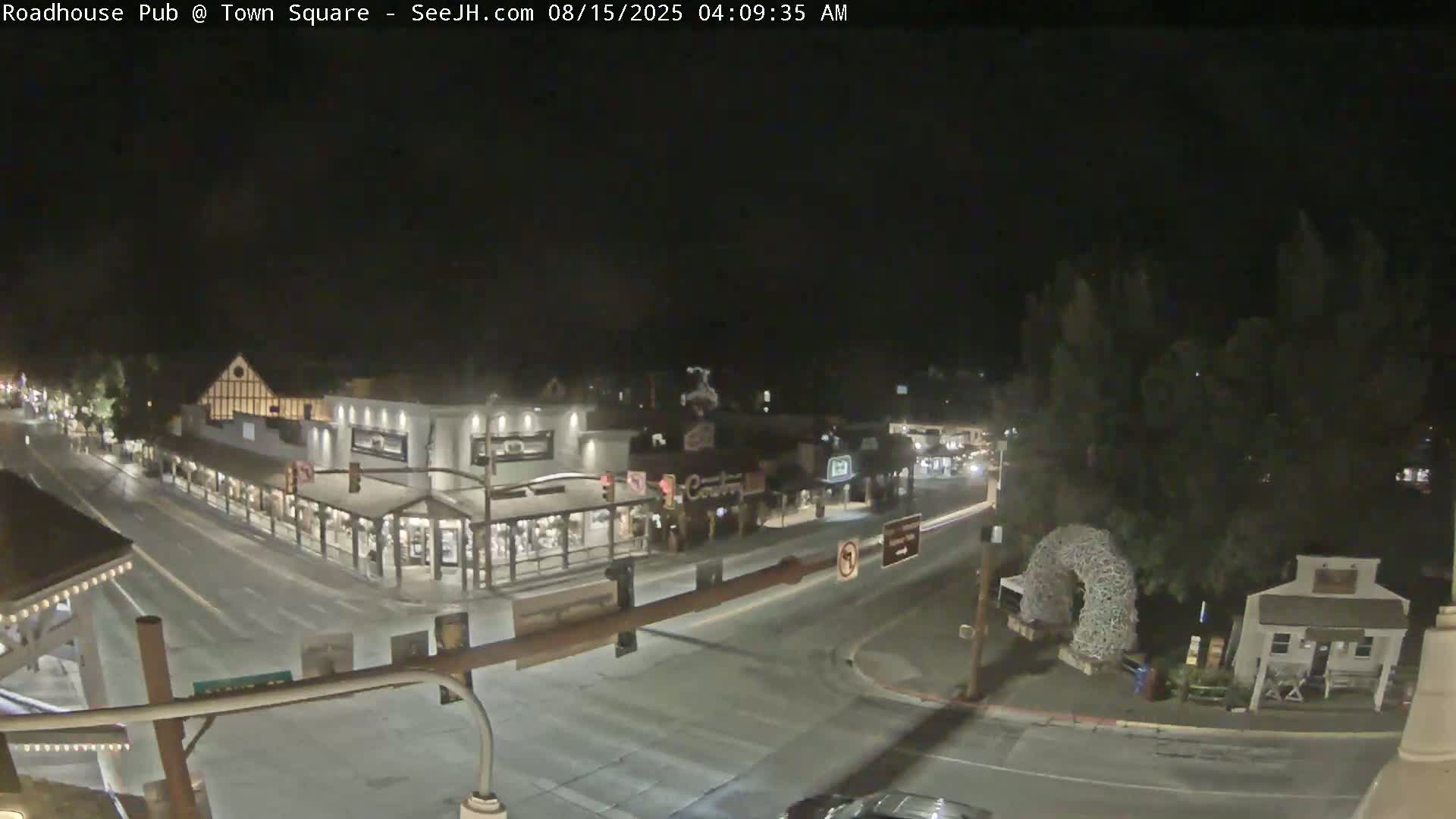A nighttime, overhead view of a town square with several buildings illuminated, a few cars on the road, and some trees.