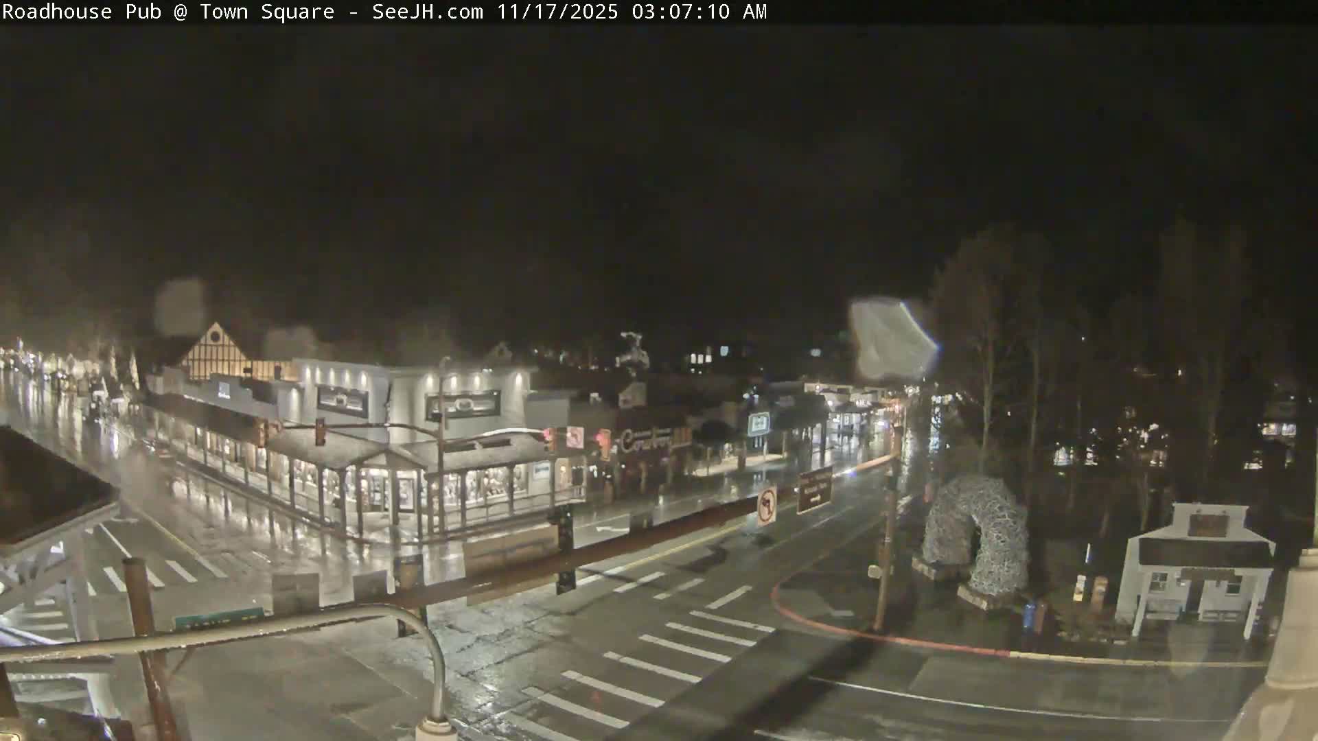 An overhead nighttime view reveals a town square with wet, reflective streets and sidewalks, lined with illuminated buildings and bare trees, under a dark, possibly misty sky.