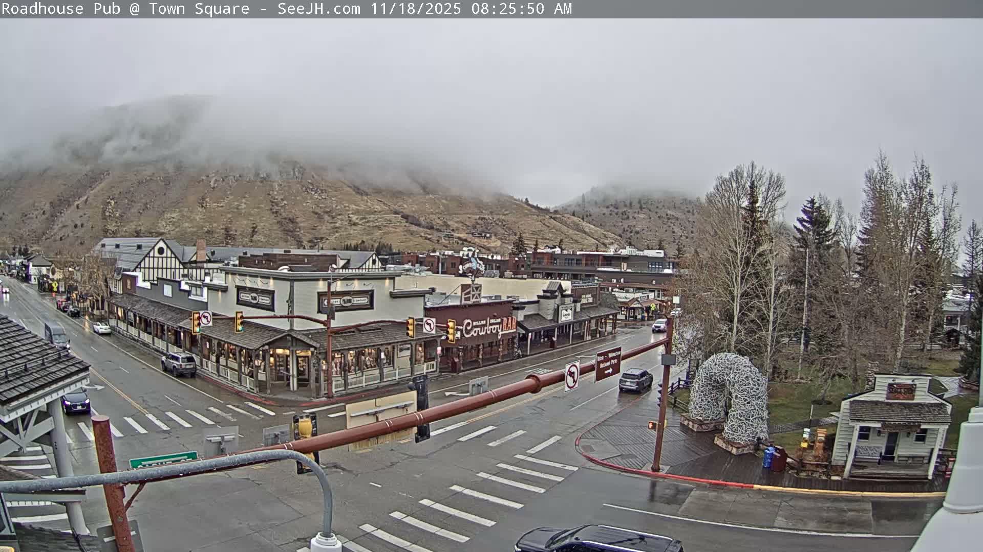 An overhead nighttime view reveals a town square with wet, reflective streets and sidewalks, lined with illuminated buildings and bare trees, under a dark, possibly misty sky.