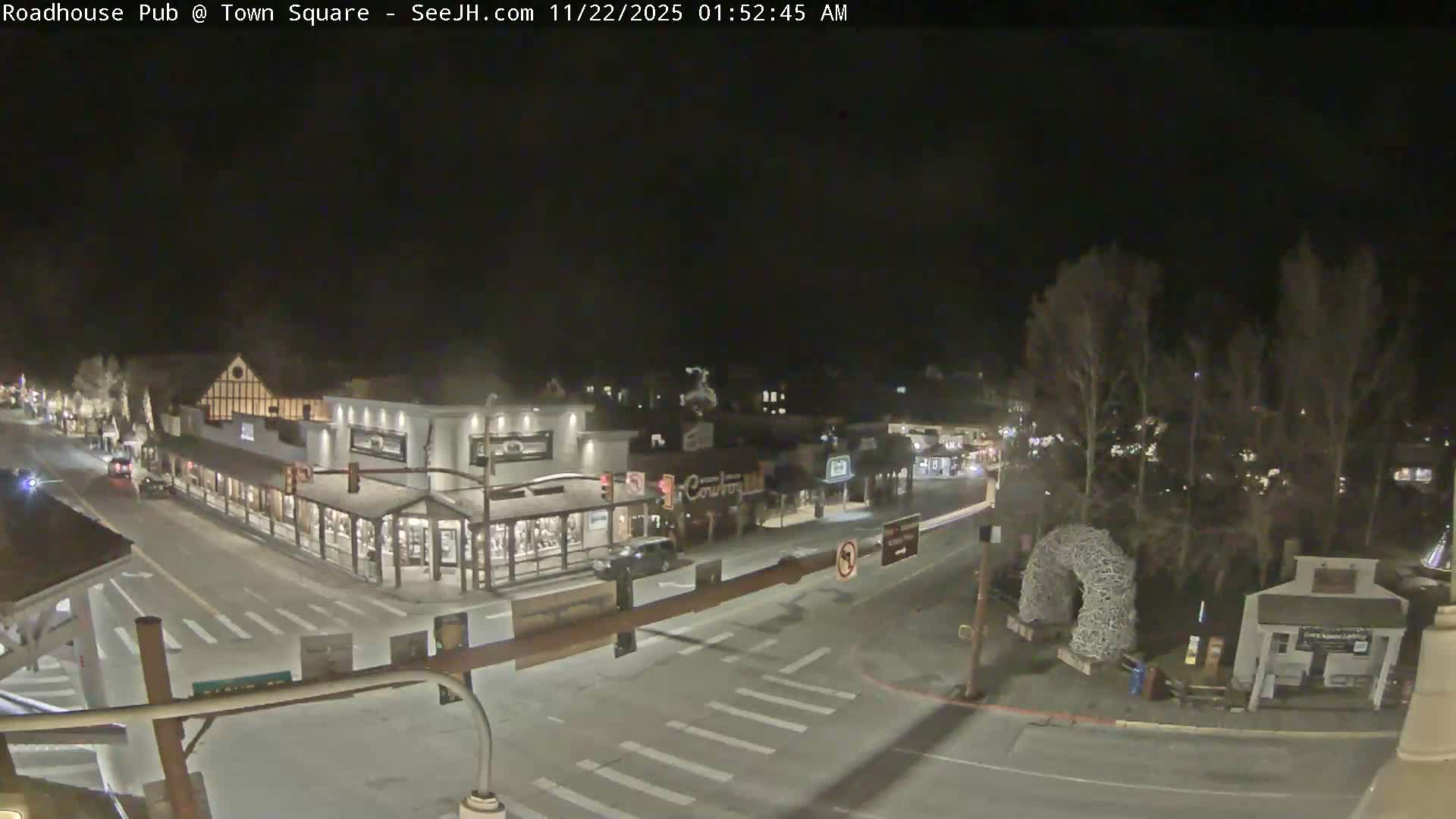 A clear night view of a town square reveals illuminated buildings and shops along a street with several cars, alongside bare trees and a unique archway sculpture on the right.