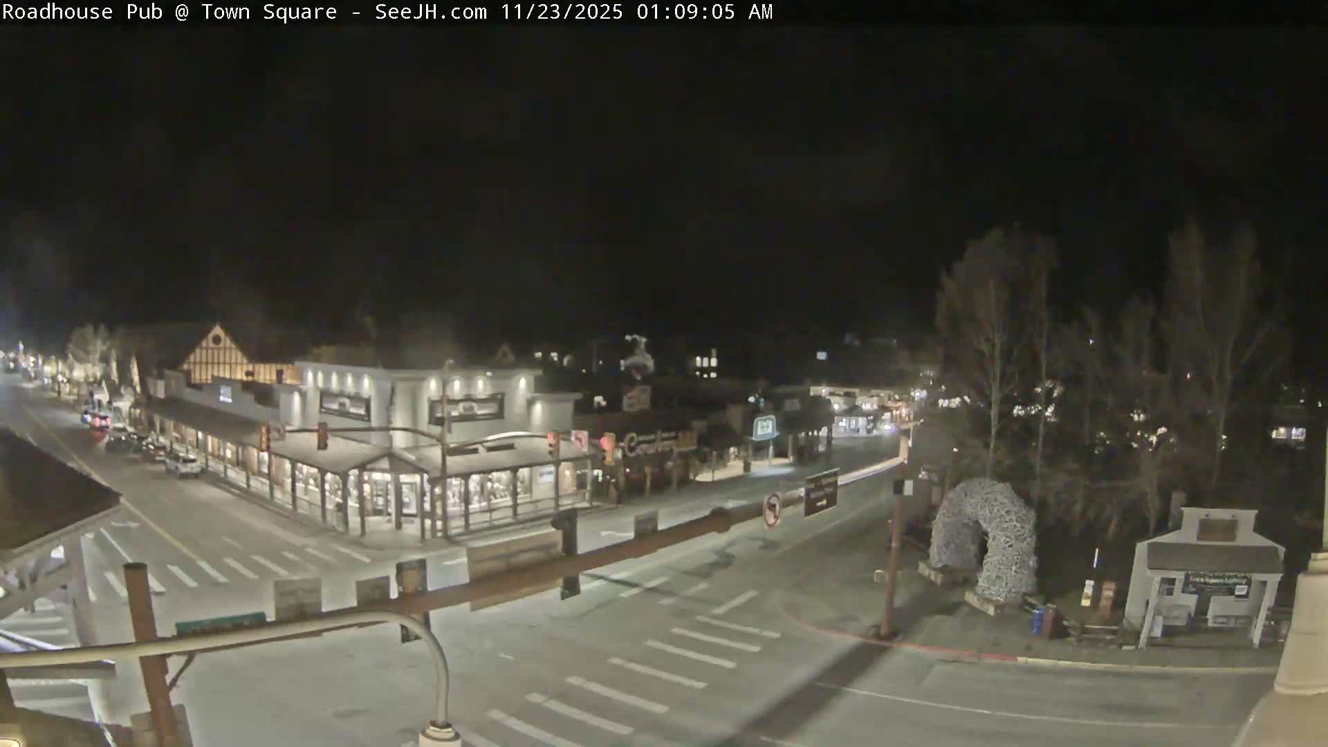 The image shows a clear night scene of a town square featuring illuminated buildings with shops, a street with a few vehicles, bare trees, and a unique arch sculpture made of interwoven material.