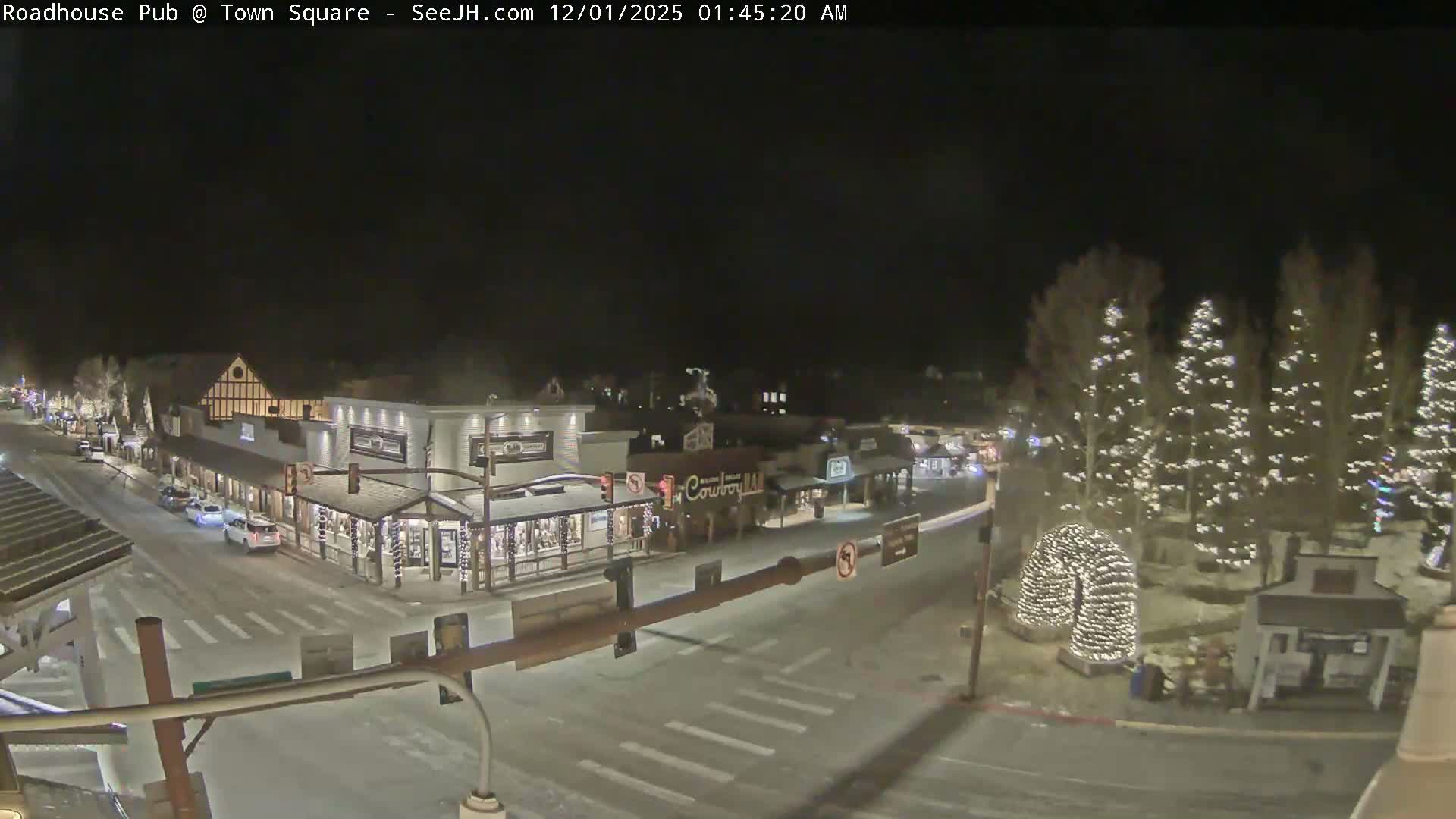 A snow-dusted town square at night is brightly illuminated by numerous string lights on trees and buildings, with a few cars on the street under a clear, cold sky.