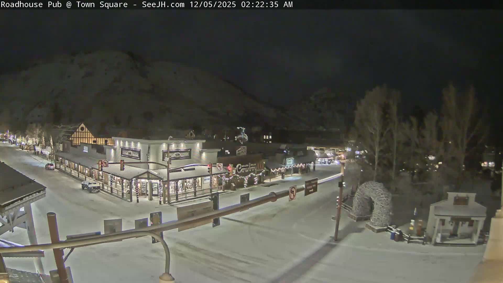 A snow-covered town street, lined with warmly lit buildings and festive decorations, is viewed at night against a backdrop of dark mountains under clear and cold conditions.