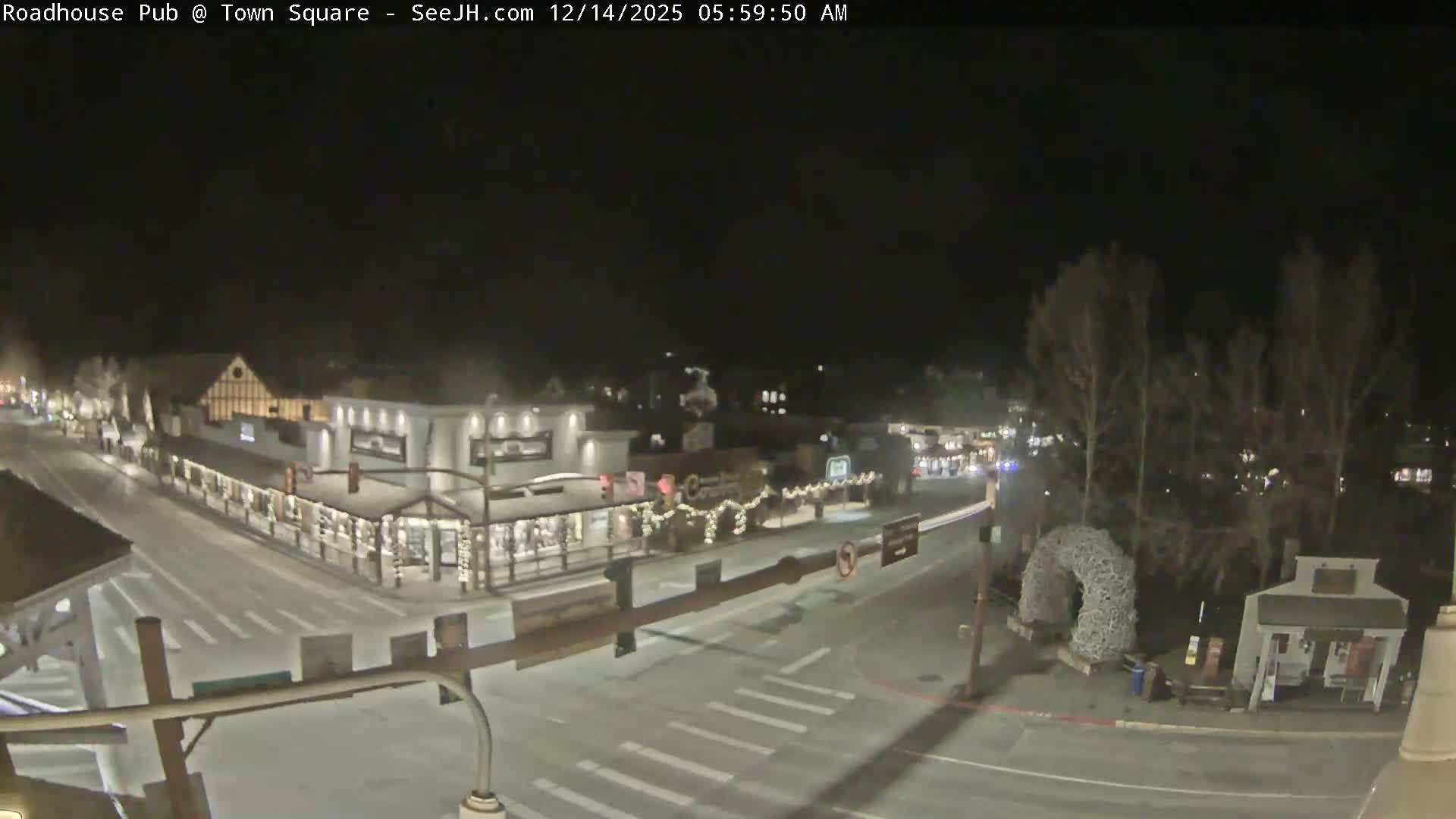 A snow-covered town street, lined with warmly lit buildings and festive decorations, is viewed at night against a backdrop of dark mountains under clear and cold conditions.