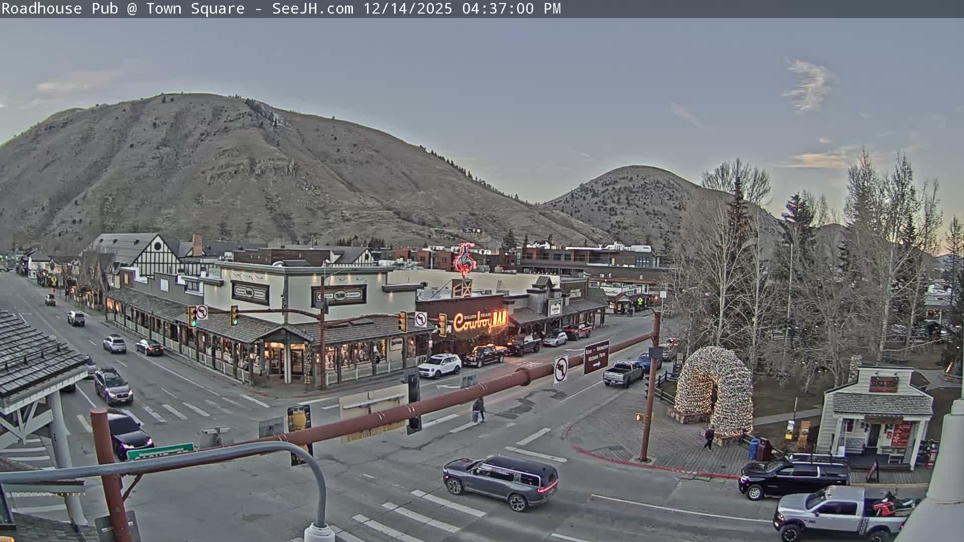 A snow-covered town street, lined with warmly lit buildings and festive decorations, is viewed at night against a backdrop of dark mountains under clear and cold conditions.
