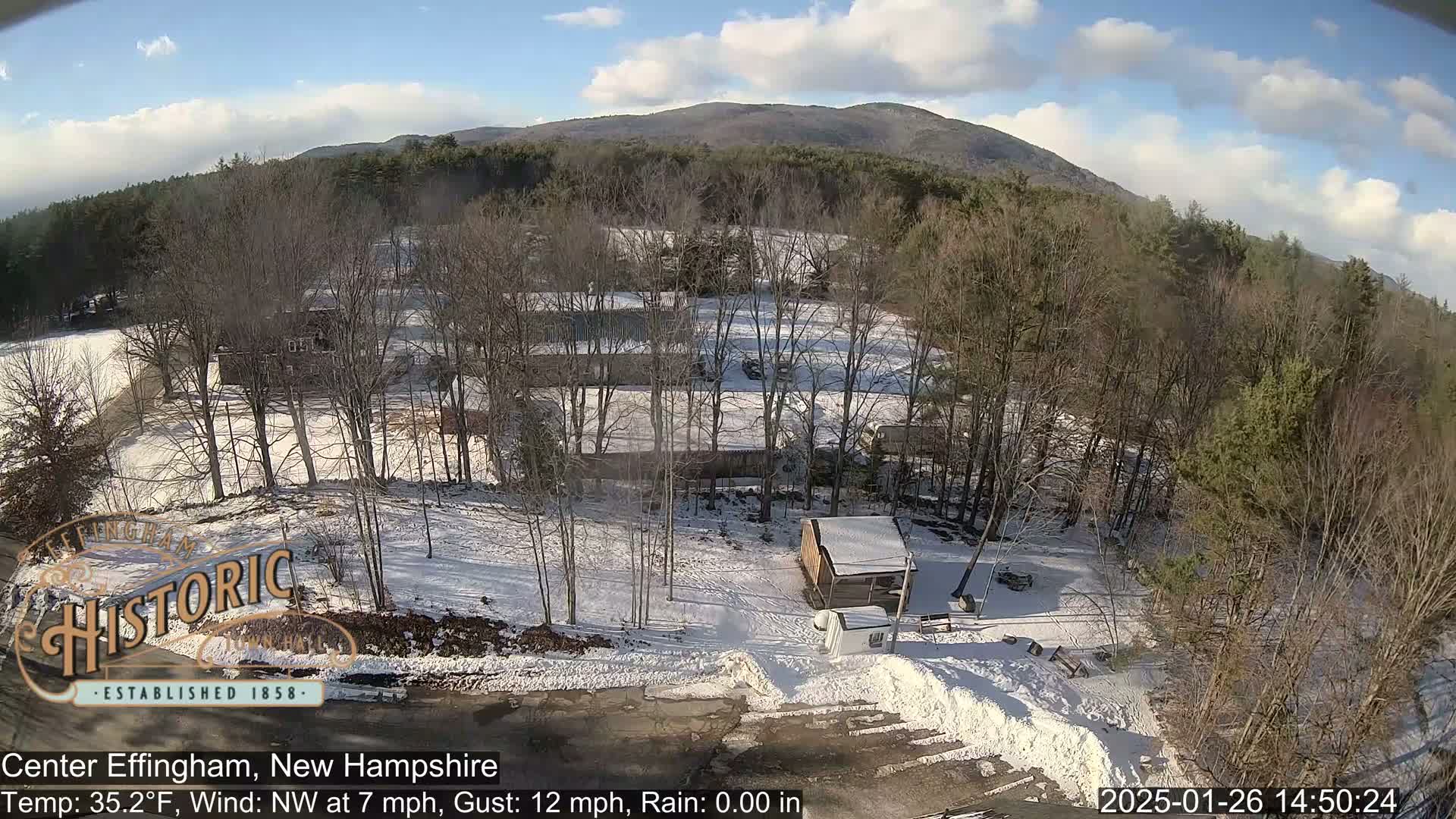 Here is a description of the image: An aerial view shows a snow-covered landscape with leafless trees, some buildings, and a small shed under a partly sunny sky.
