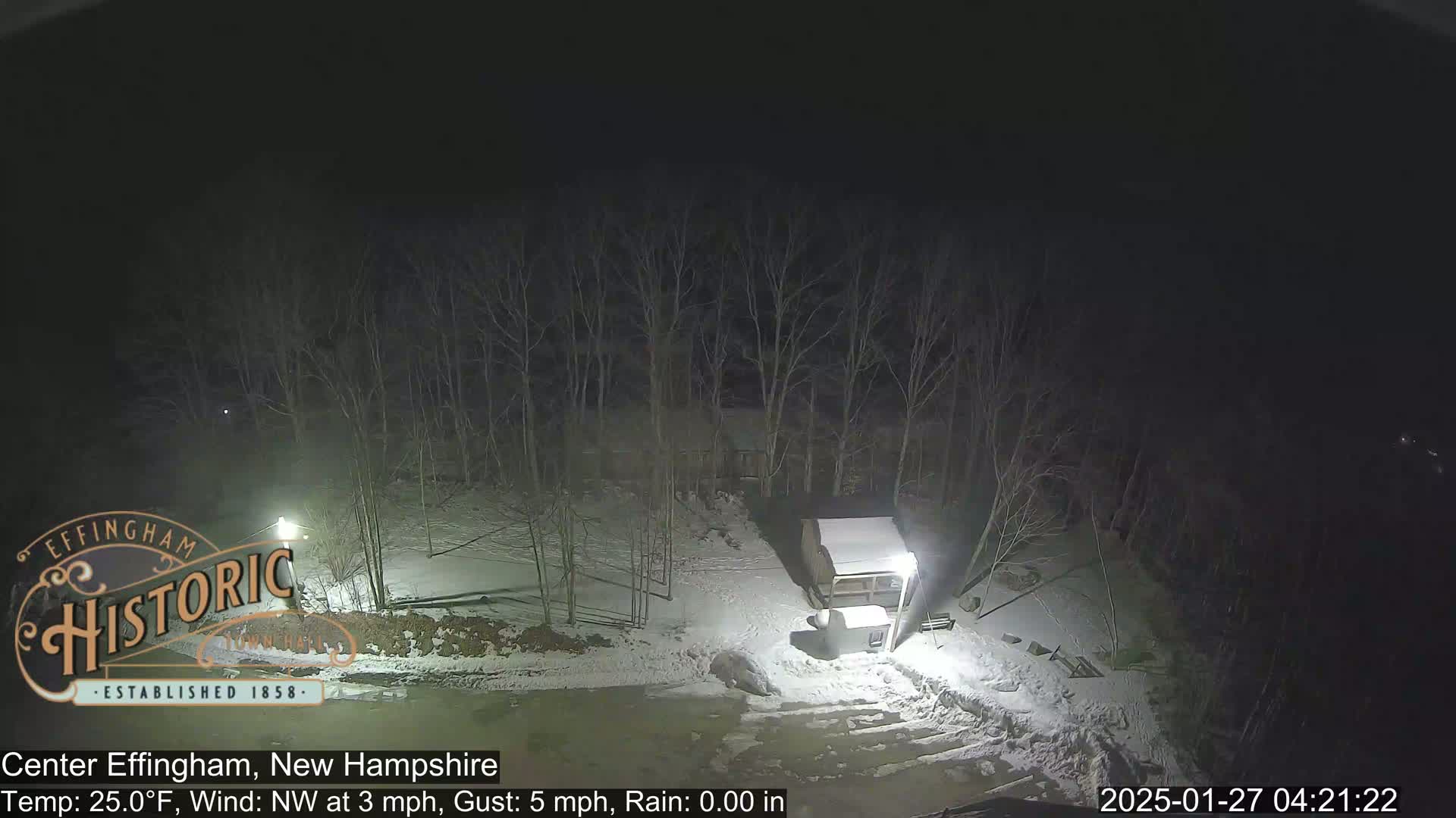 A nighttime view of a snow-covered area with a small building and bare trees under a dark sky.