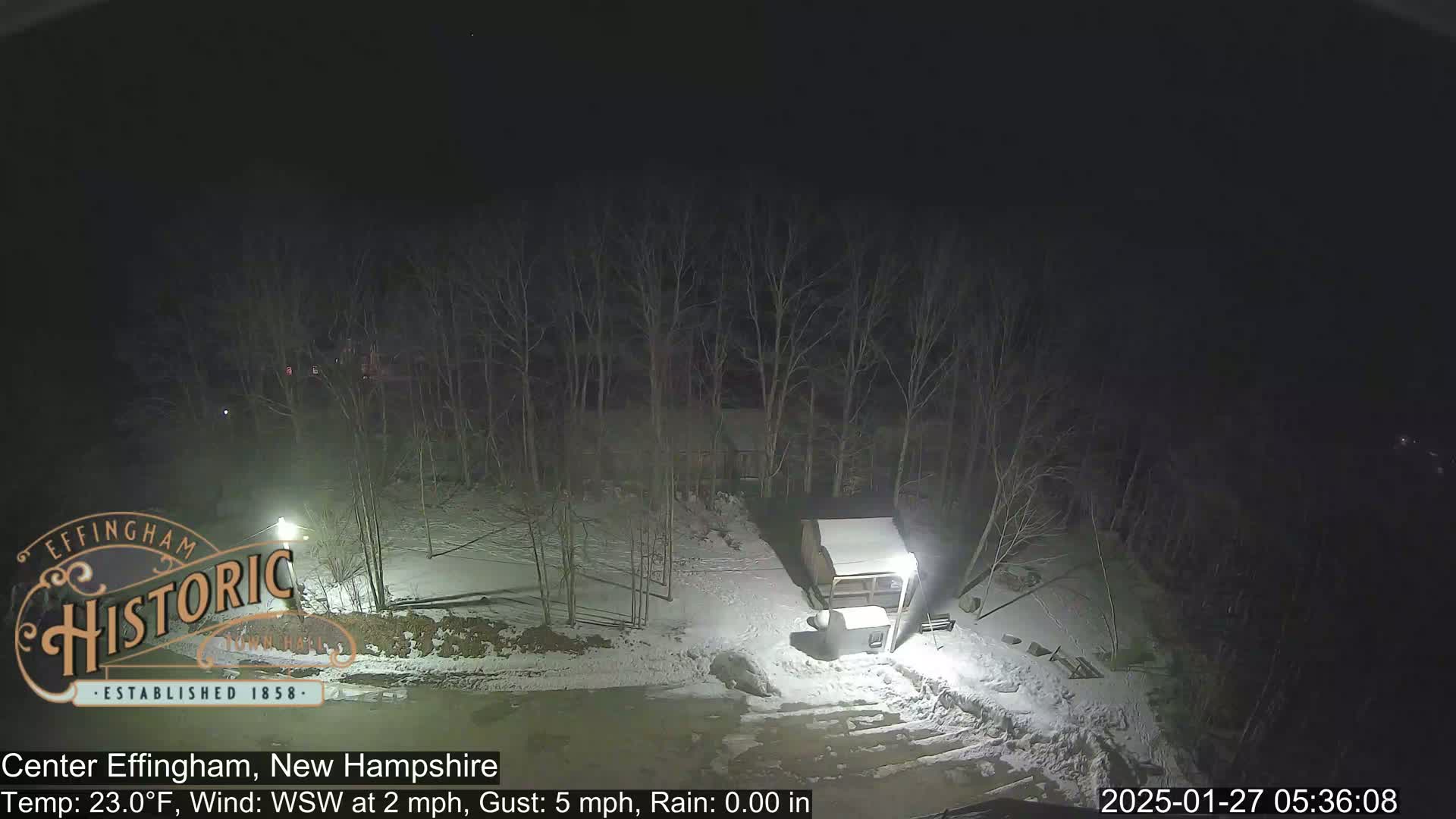 A nighttime scene of a snow-covered area with bare trees and a small building, under conditions of light rain and a temperature of 23 degrees Fahrenheit.