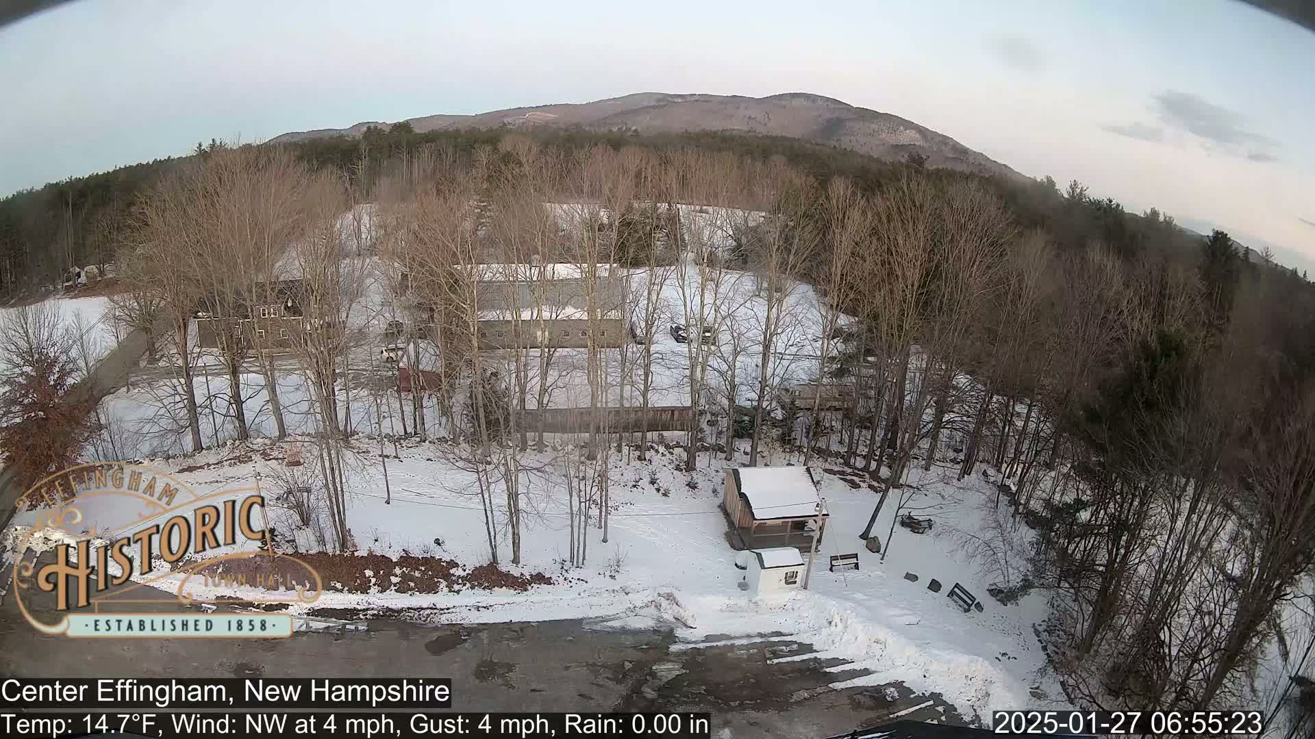 Here is a description of the image: An aerial view shows a snow-covered area with several buildings, bare trees, and a mountain in the background under a clear sky.