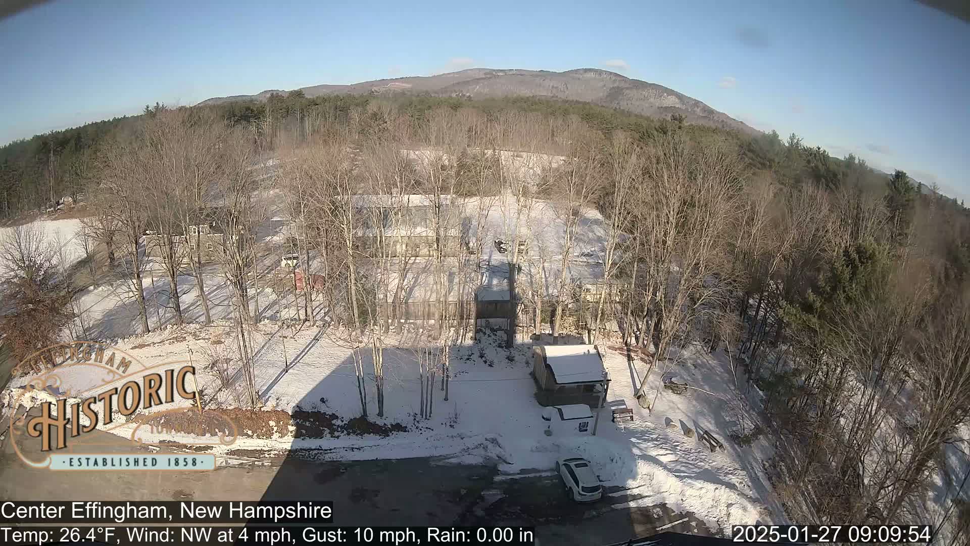 A snow-covered landscape with bare trees, several buildings, and a car is visible under a clear, sunny sky.