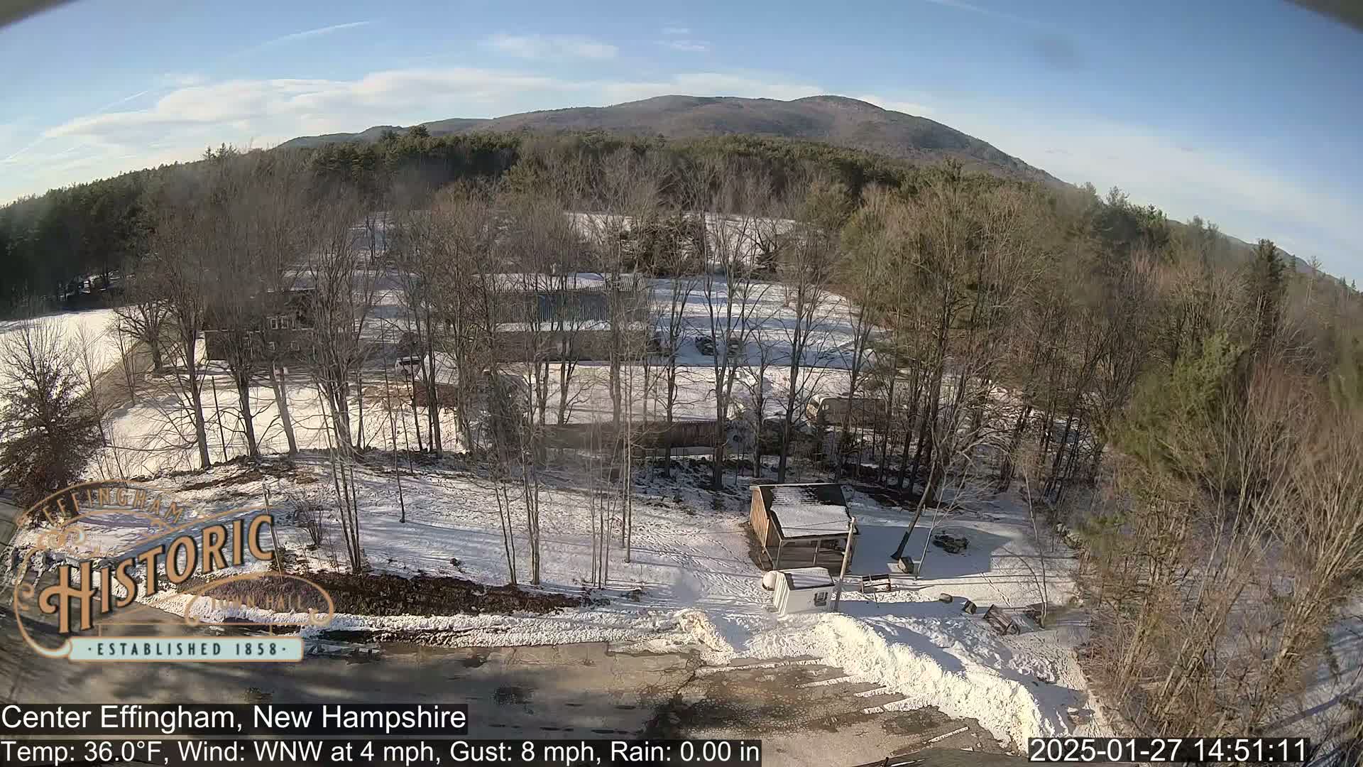 A snow-covered landscape with bare trees and a few buildings, under a sunny sky, is viewed from above.