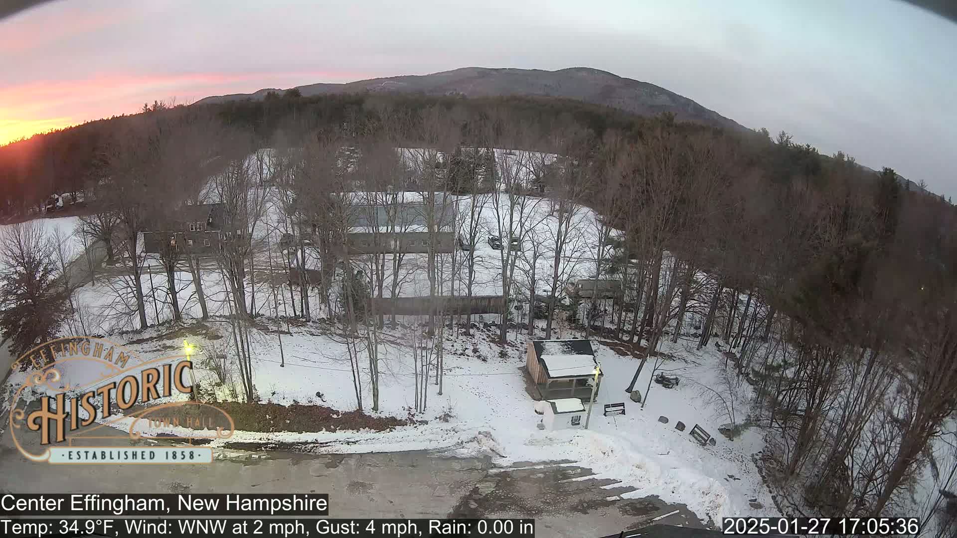 A snow-covered landscape at sunset, featuring several buildings and bare trees, with a mountain in the background.
