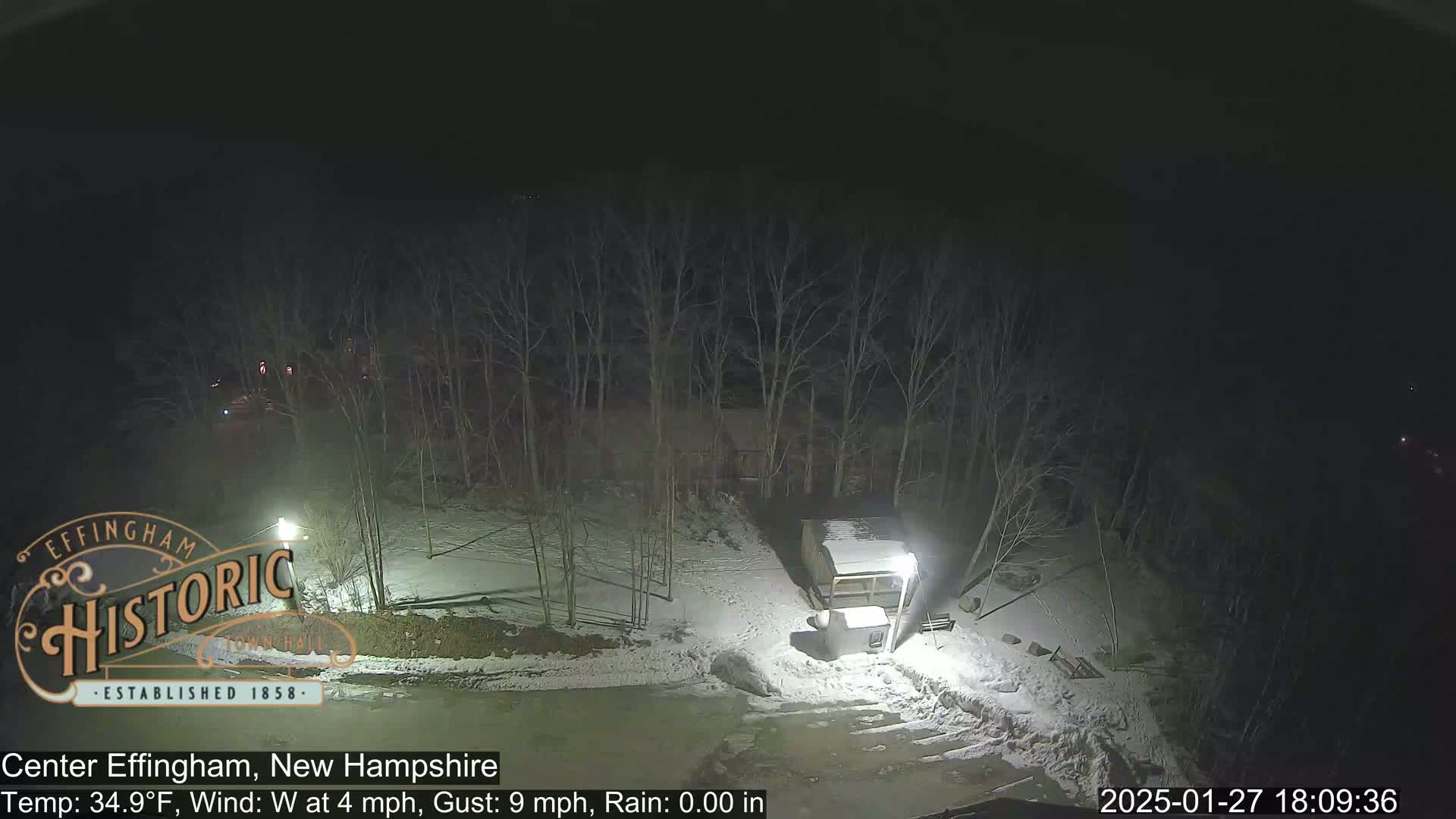 A nighttime view of a snow-covered area with bare trees and a small structure, under a dark, clear sky with a westerly wind.