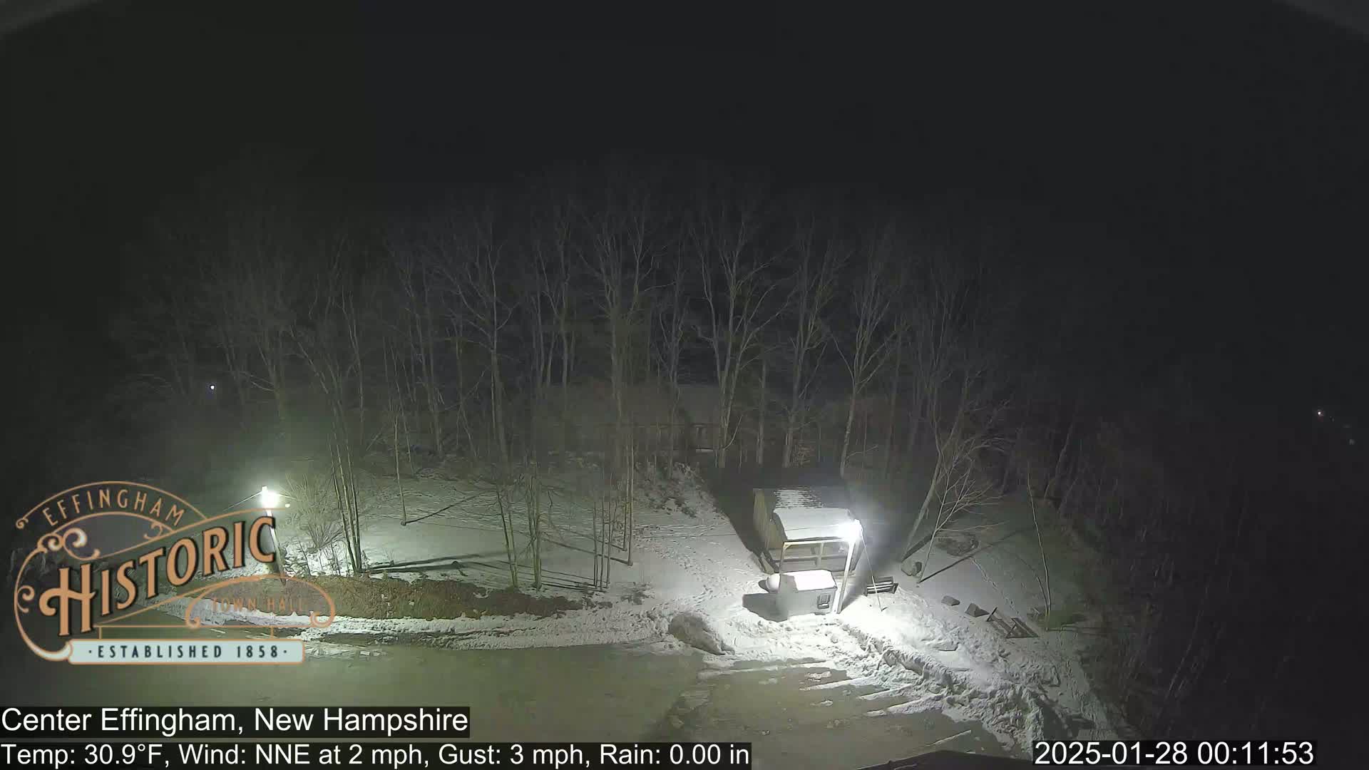 A nighttime, snow-covered scene shows bare trees and a small structure illuminated by lights, with a temperature of 30.9°F and a light north-northeast wind.