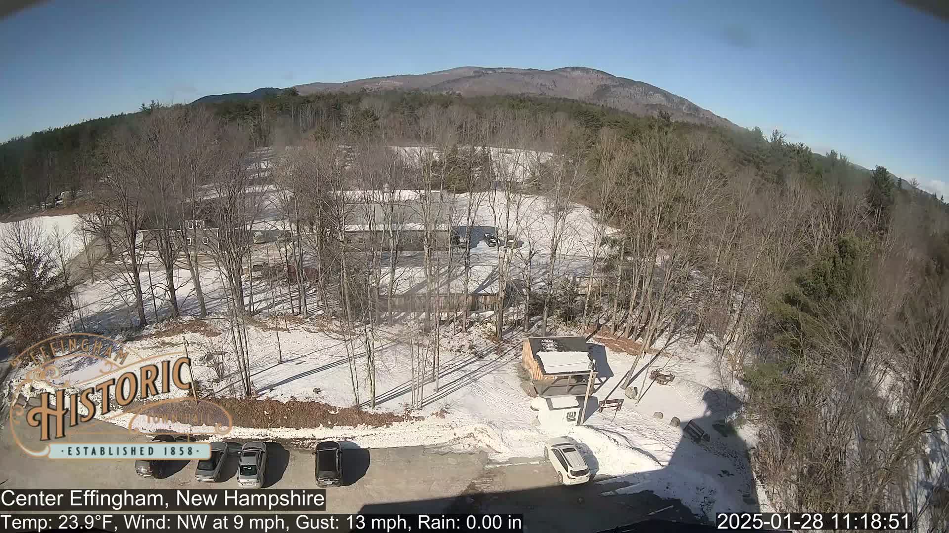 A snow-covered parking lot with several cars, a small building, and bare trees, situated below a mountain range under a clear, sunny sky.