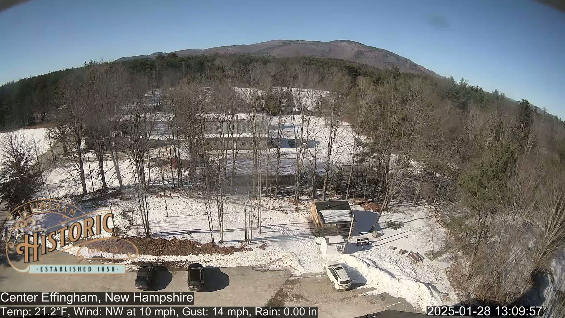 A snow-covered parking lot with two cars and a small building, surrounded by bare trees and a mountain in the background under a clear, sunny sky.