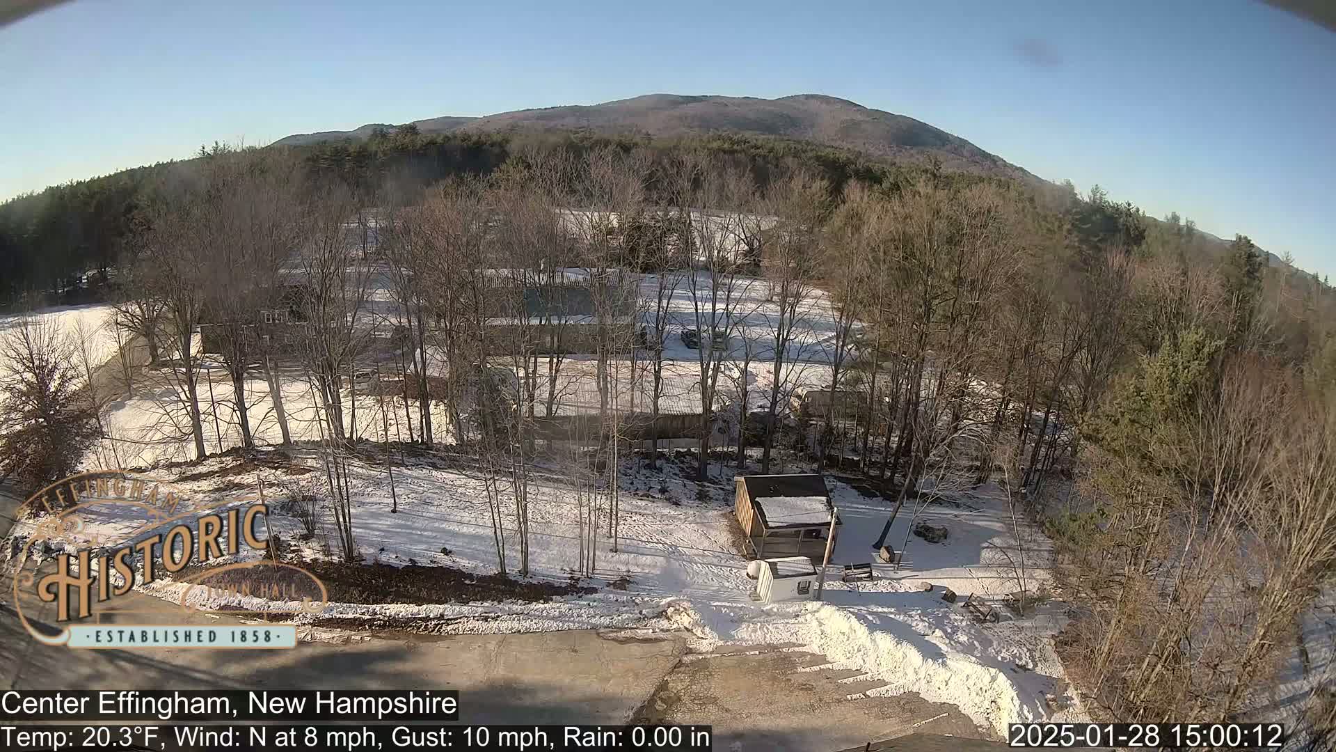 A snow-covered landscape with leafless trees, several buildings, and a small shed is visible under a clear, sunny sky.