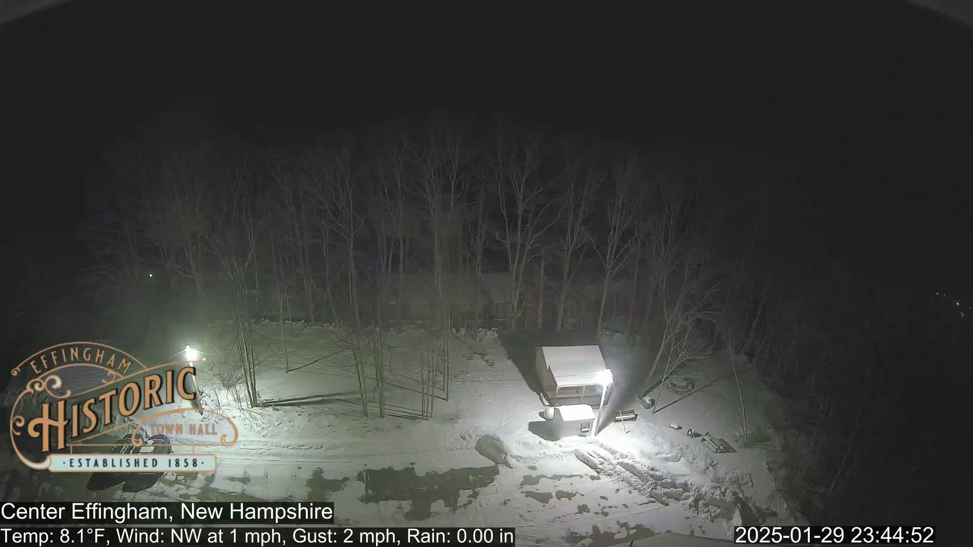 A nighttime, snow-covered scene shows bare trees, a small shed, and a light post under a dark sky with a northwest wind.