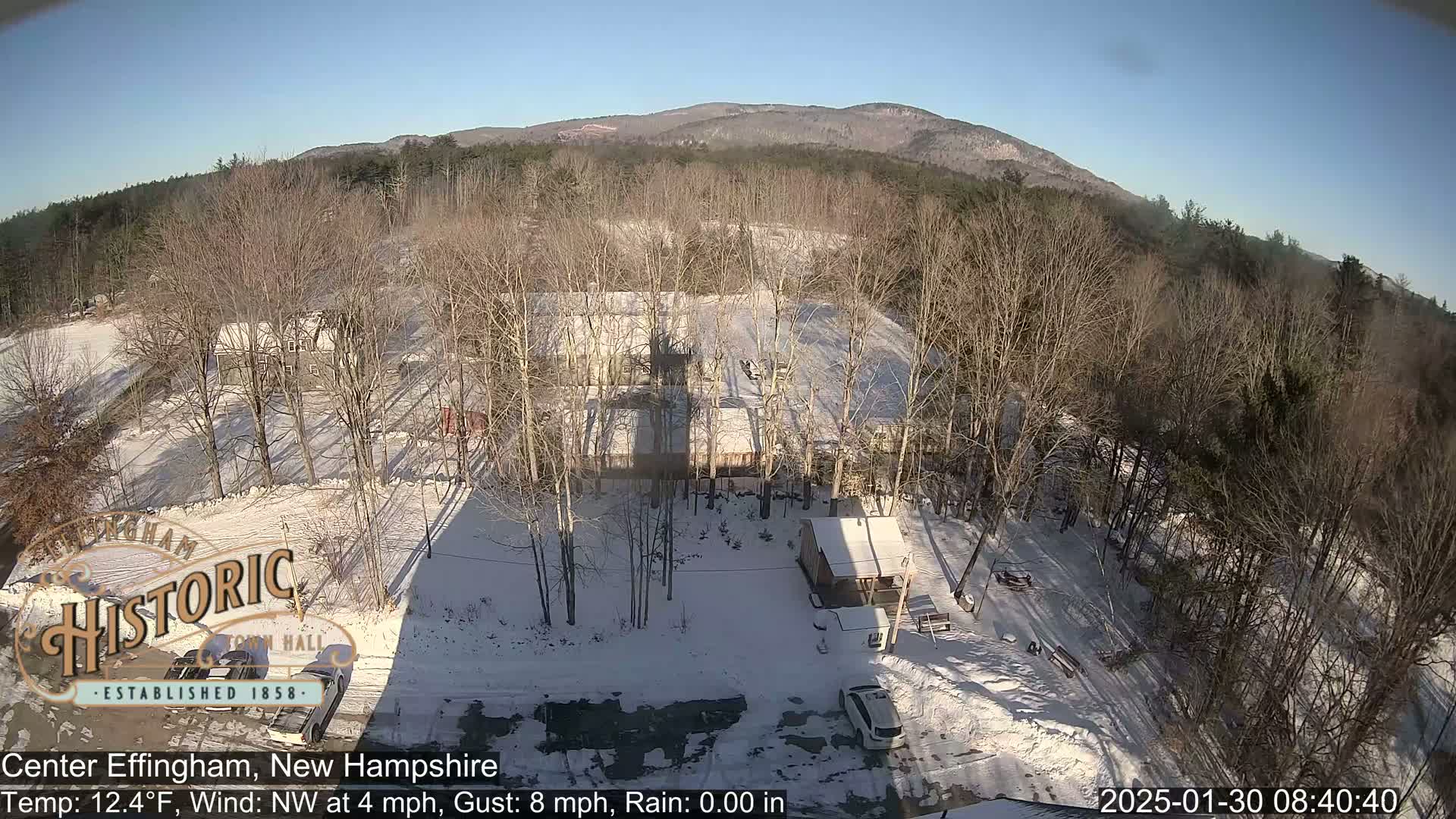 A snow-covered landscape with bare trees and a building, viewed from above under a clear, sunny sky.