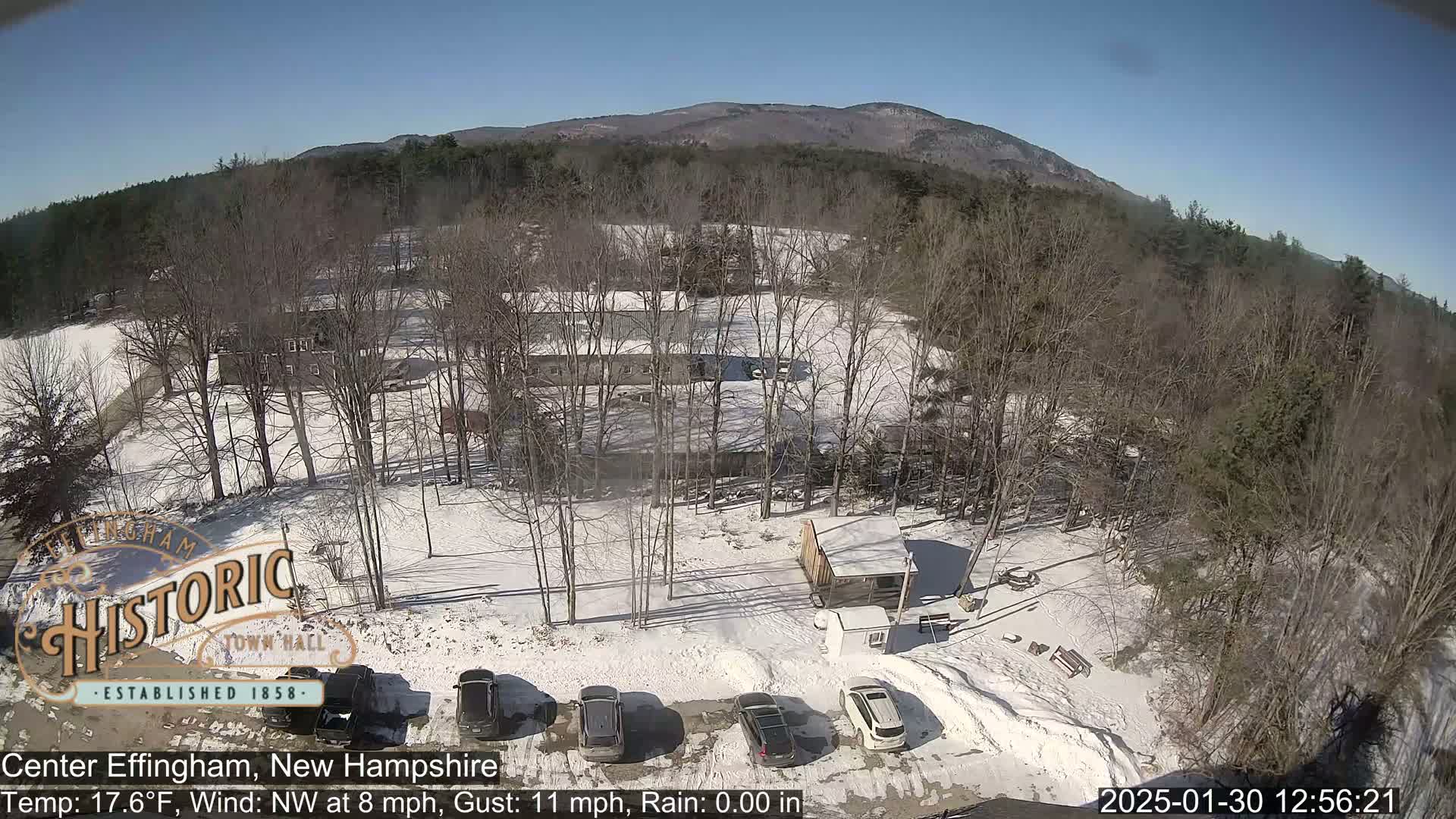 A snow-covered parking lot with several cars, a small building, and bare trees surrounds a larger building, all set against a backdrop of a distant mountain range under a clear, sunny sky.